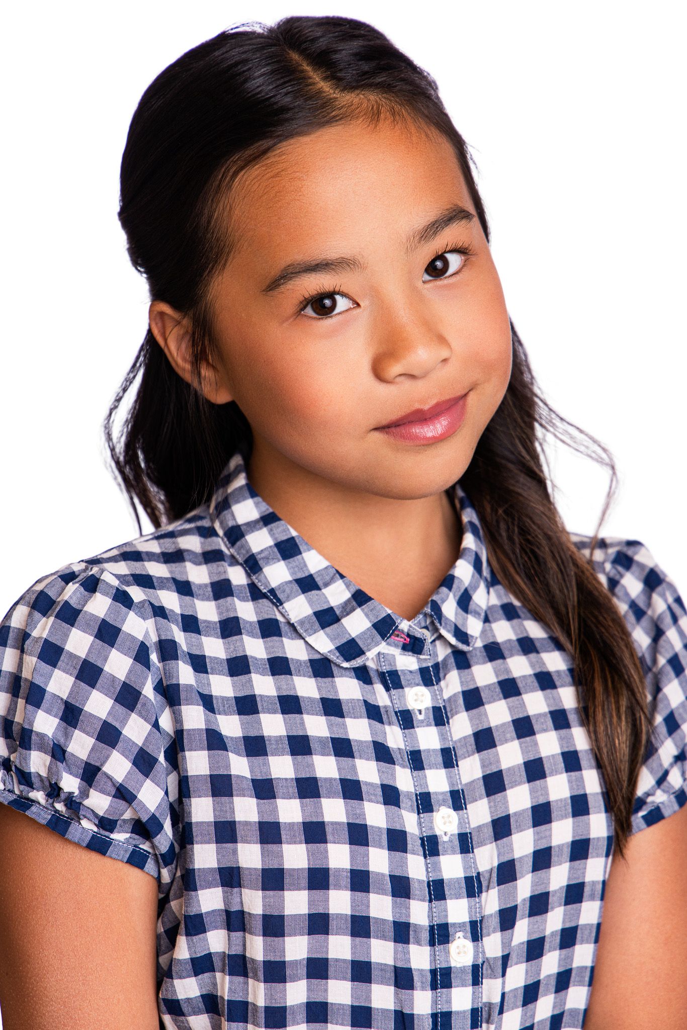 Young Asian girl in blue gingham checkered shirt posing against white background with hair pulled back.