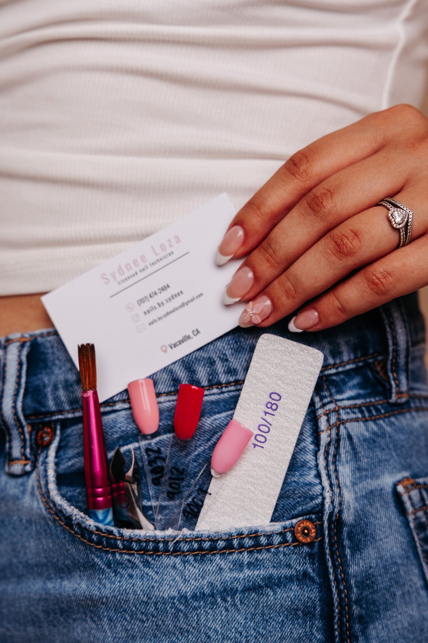 Nail technician holding business card over jeans pocket filled with nail polish, nail tips, and a nail file.