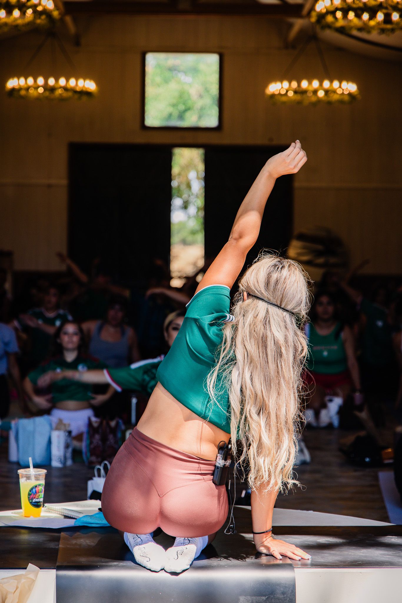 Blonde woman in green top and pink leggings kneeling on stage with arm raised during yoga class.