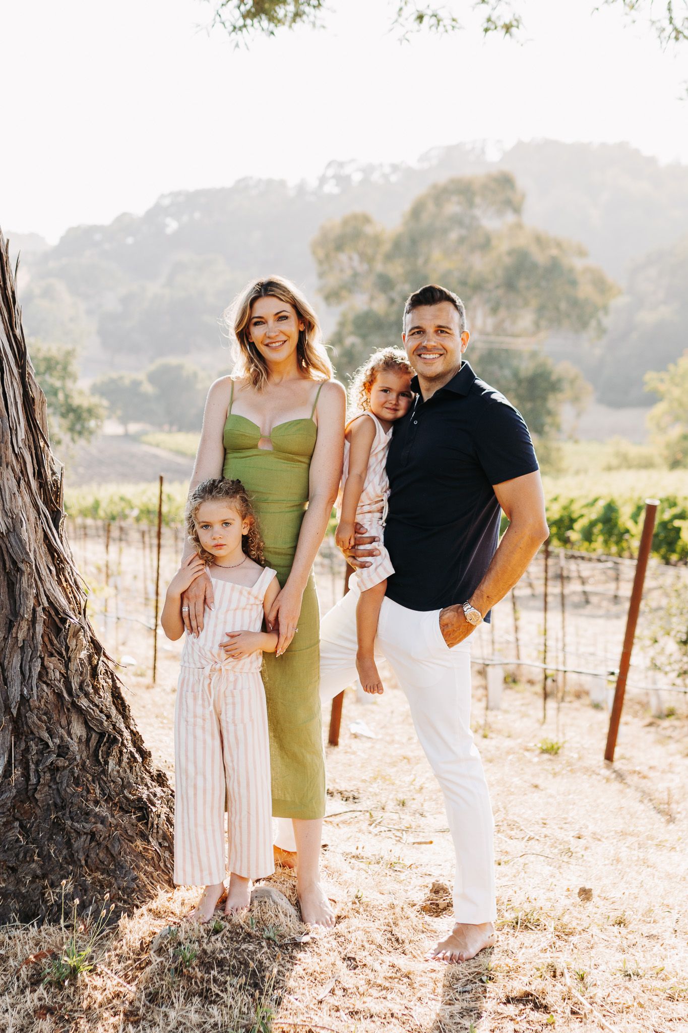 Family poses together in casual attire among vineyard rows during golden hour sunset photography session.