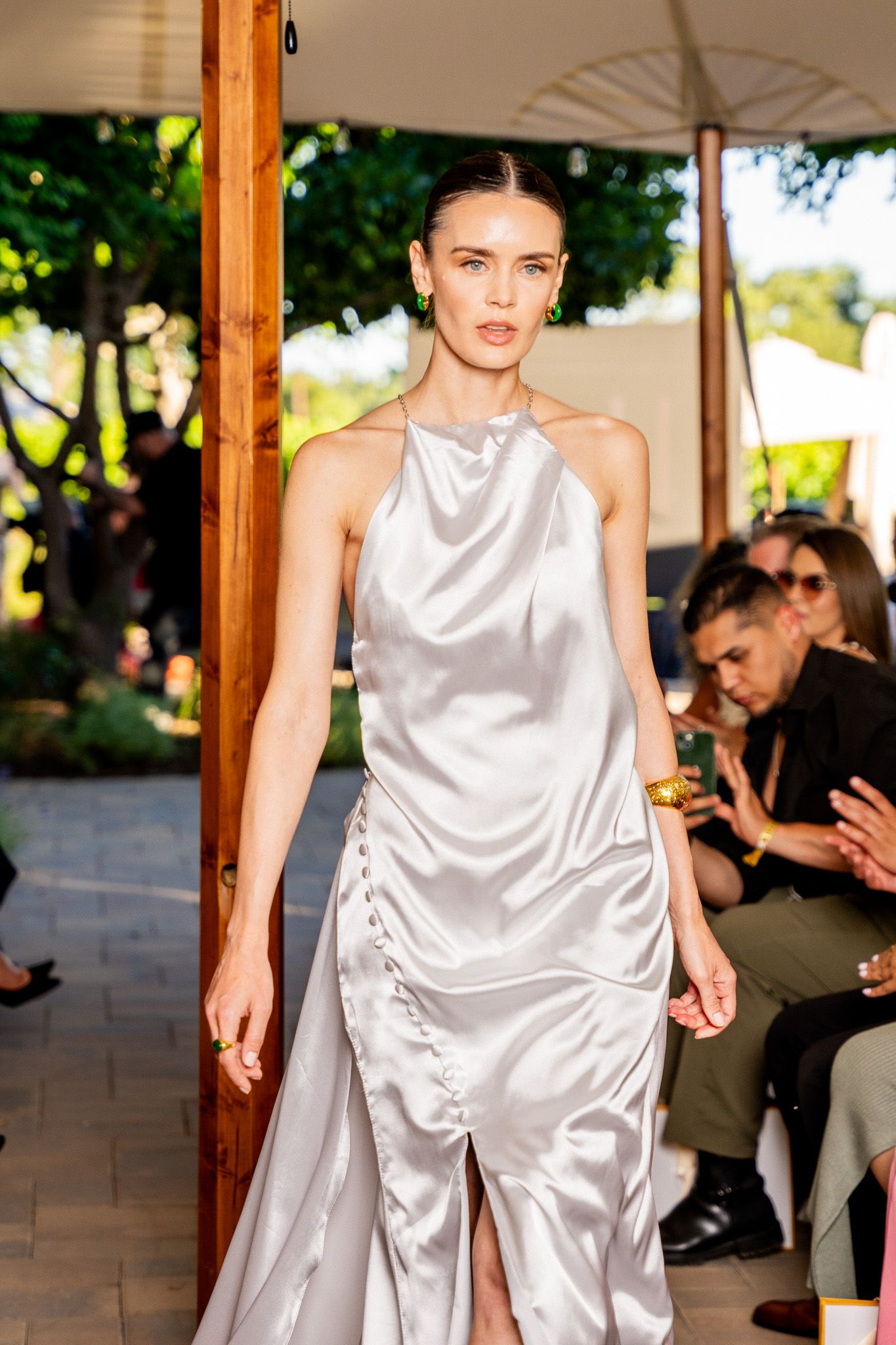 Model walks runway in flowing white satin evening gown during outdoor fashion show.
