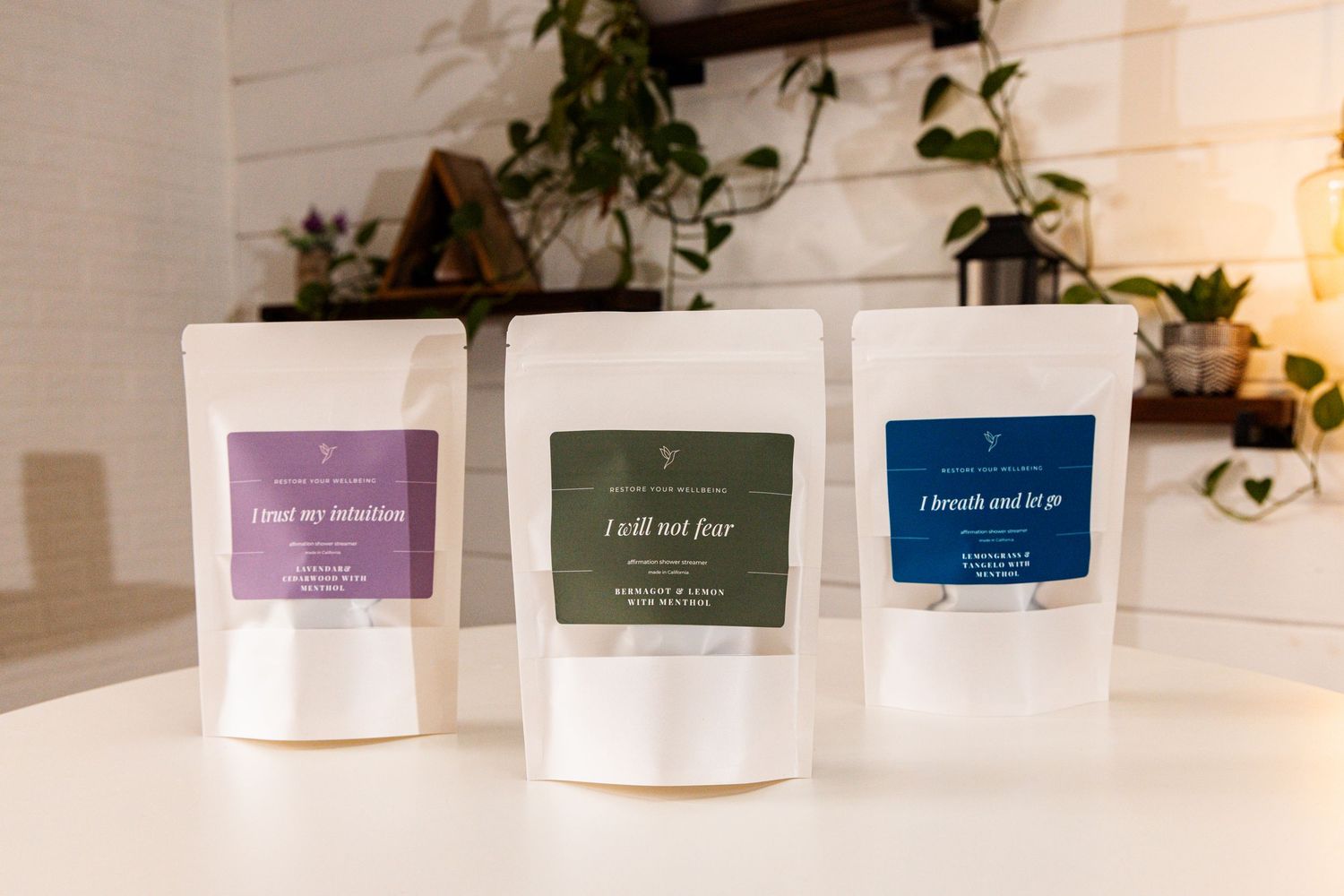 Three white pouches of loose leaf tea with purple, green, and blue labels displayed on a white counter with ivy decor.