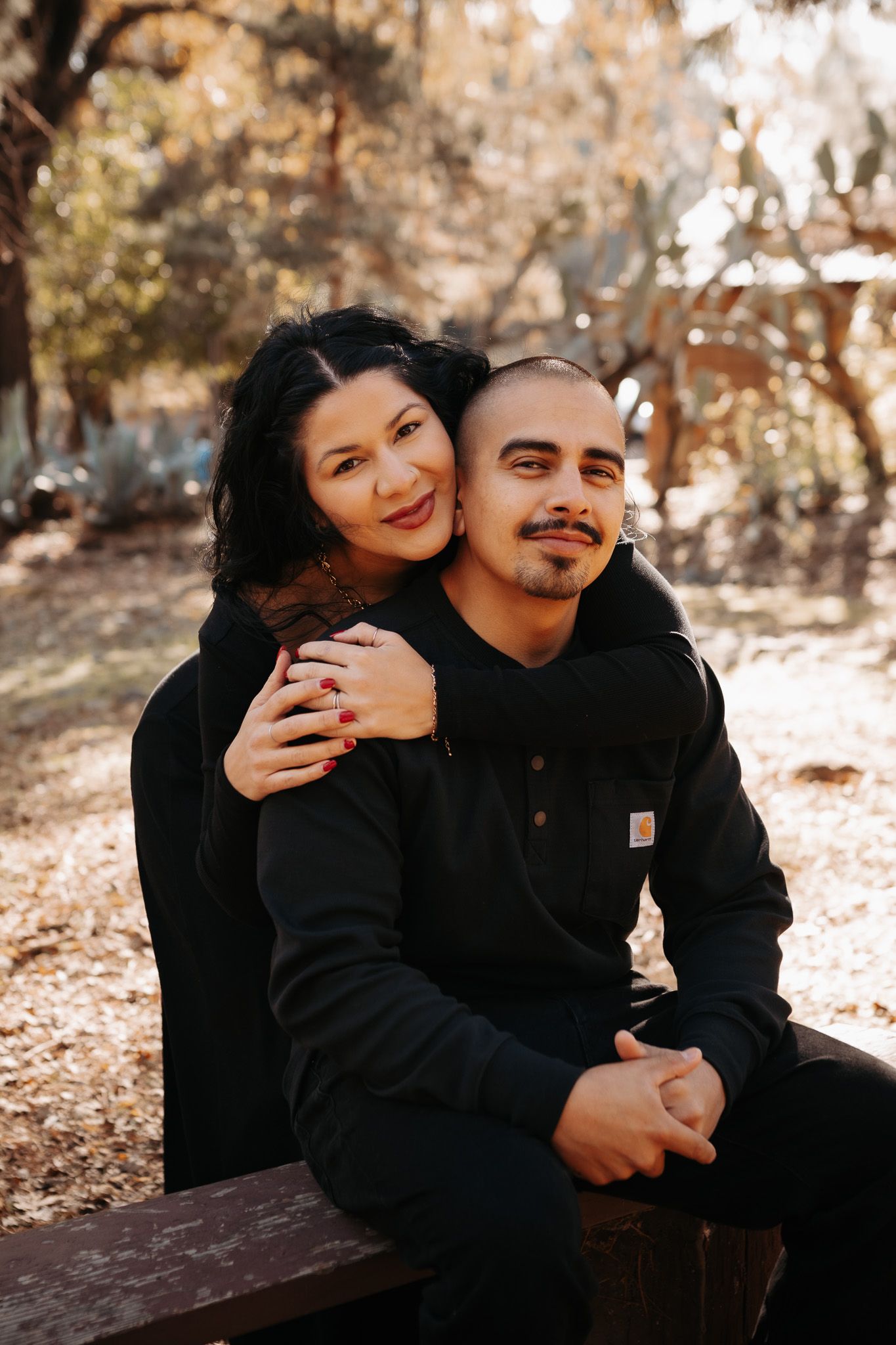 A couple wearing black outfits shares intimate moments while sitting outdoors on a wooden bench in autumn lighting.