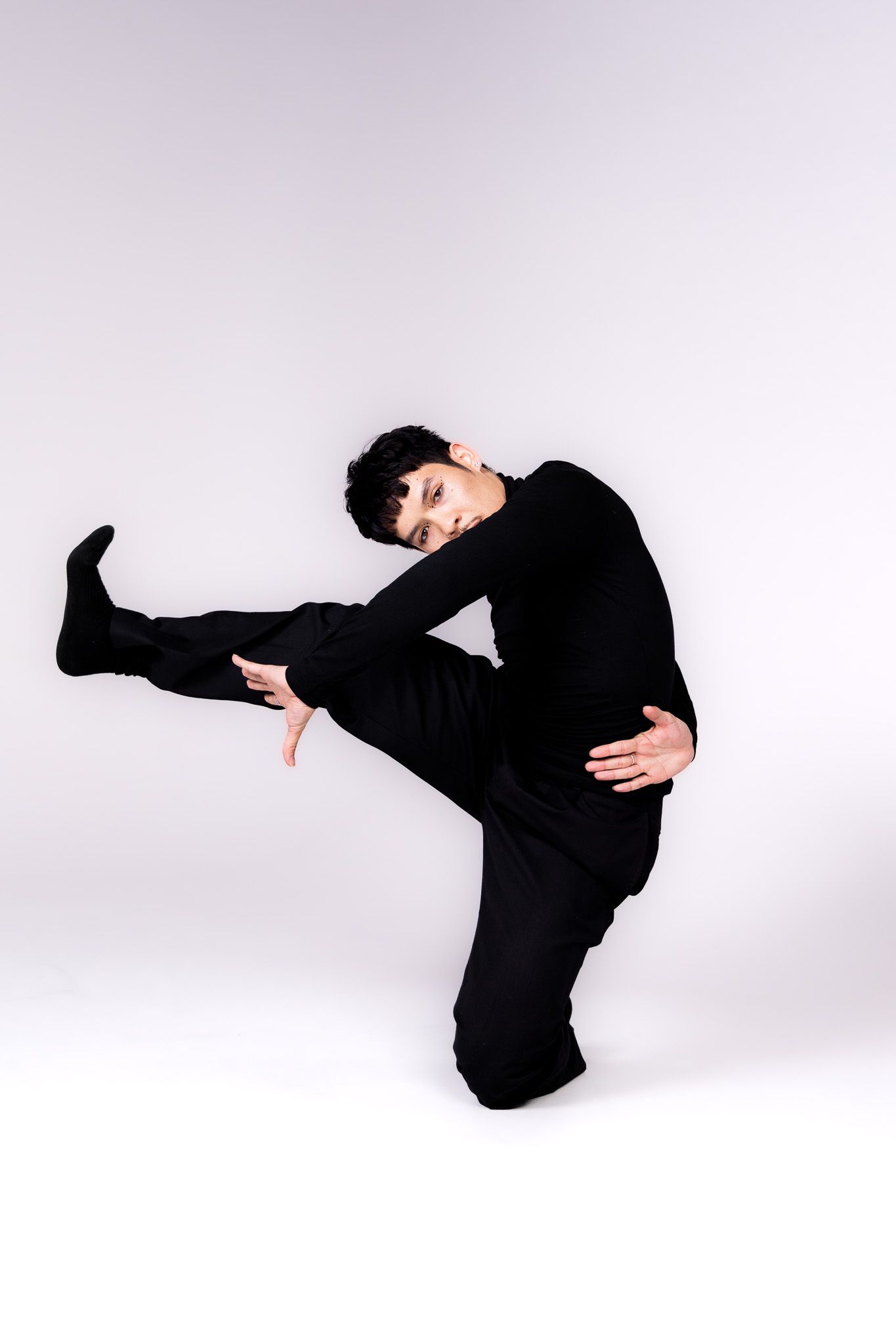 Modern dancer in black performs expressive side kick pose in studio setting.