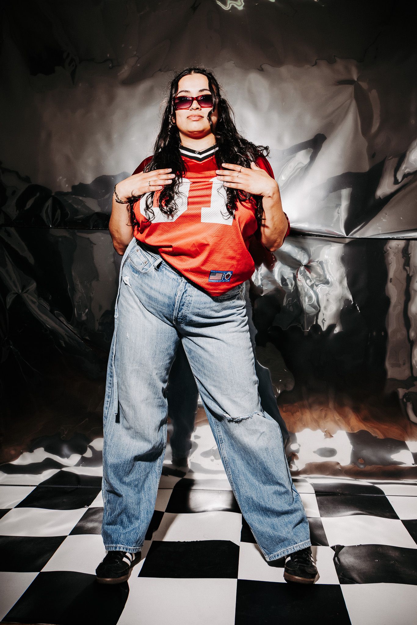 Person in baggy jeans and red top strikes pose on checkered floor against metallic backdrop with sunglasses on.
