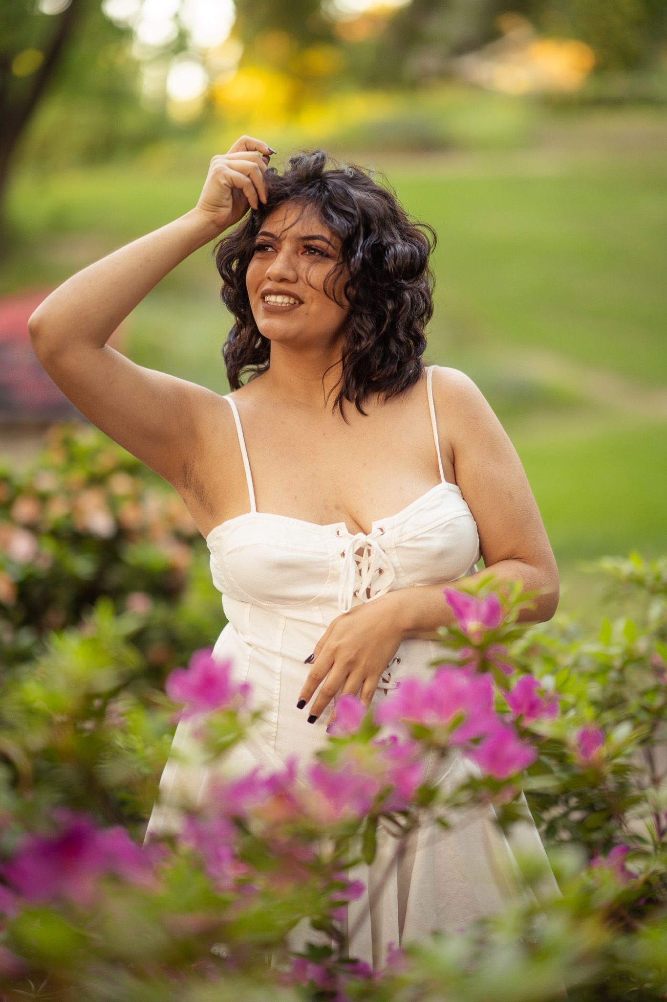 Joyful figure in white dress poses playfully amid blooming pink flowers in a garden during golden hour lighting.