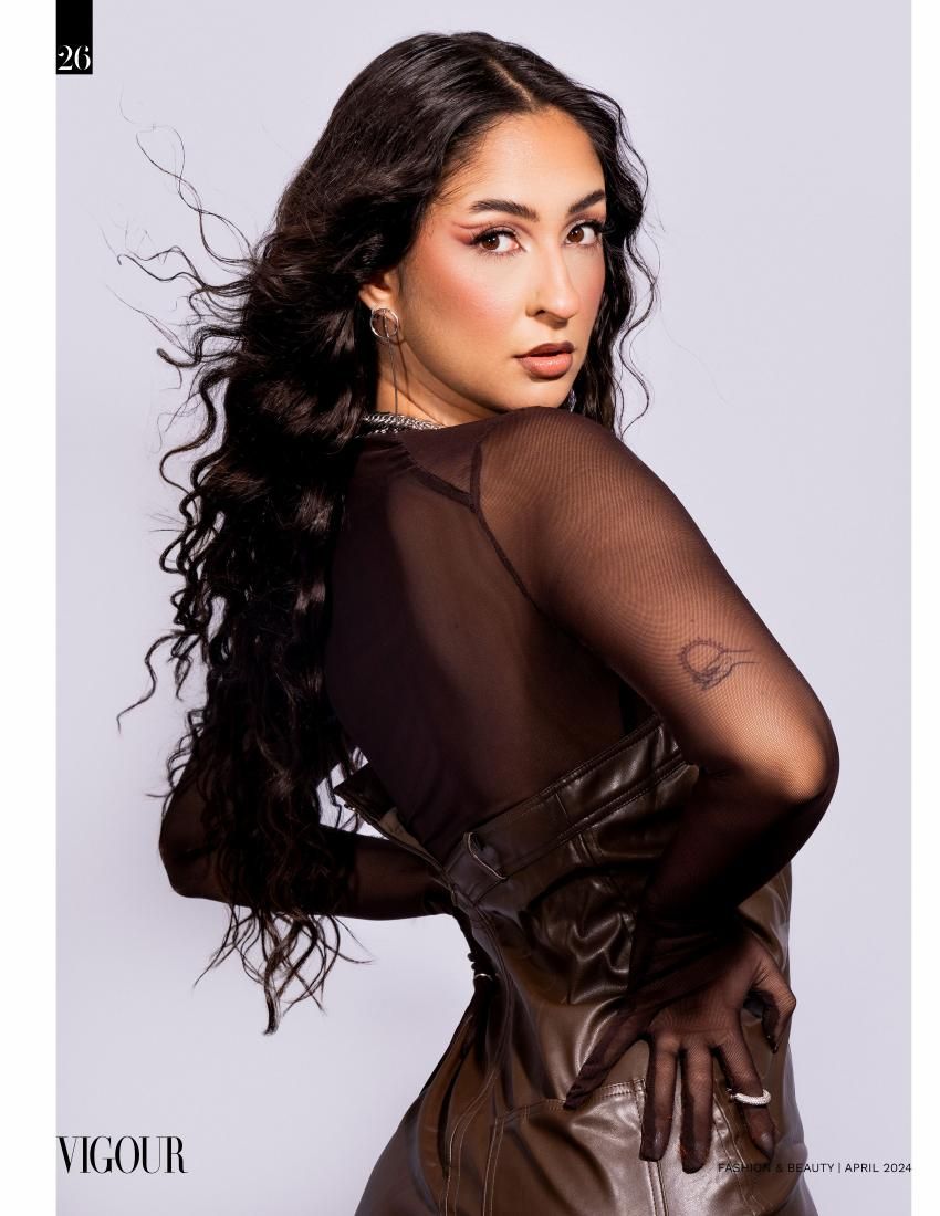 Model with long dark wavy hair poses in sheer brown garment looking over shoulder in studio portrait.