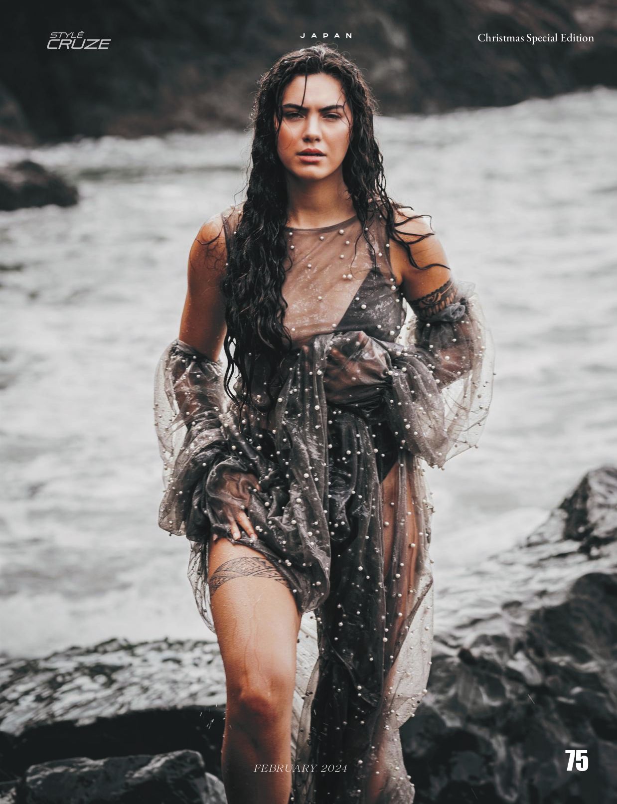 Model poses on rocky beach in sheer flowing dress during dramatic black and white photoshoot.