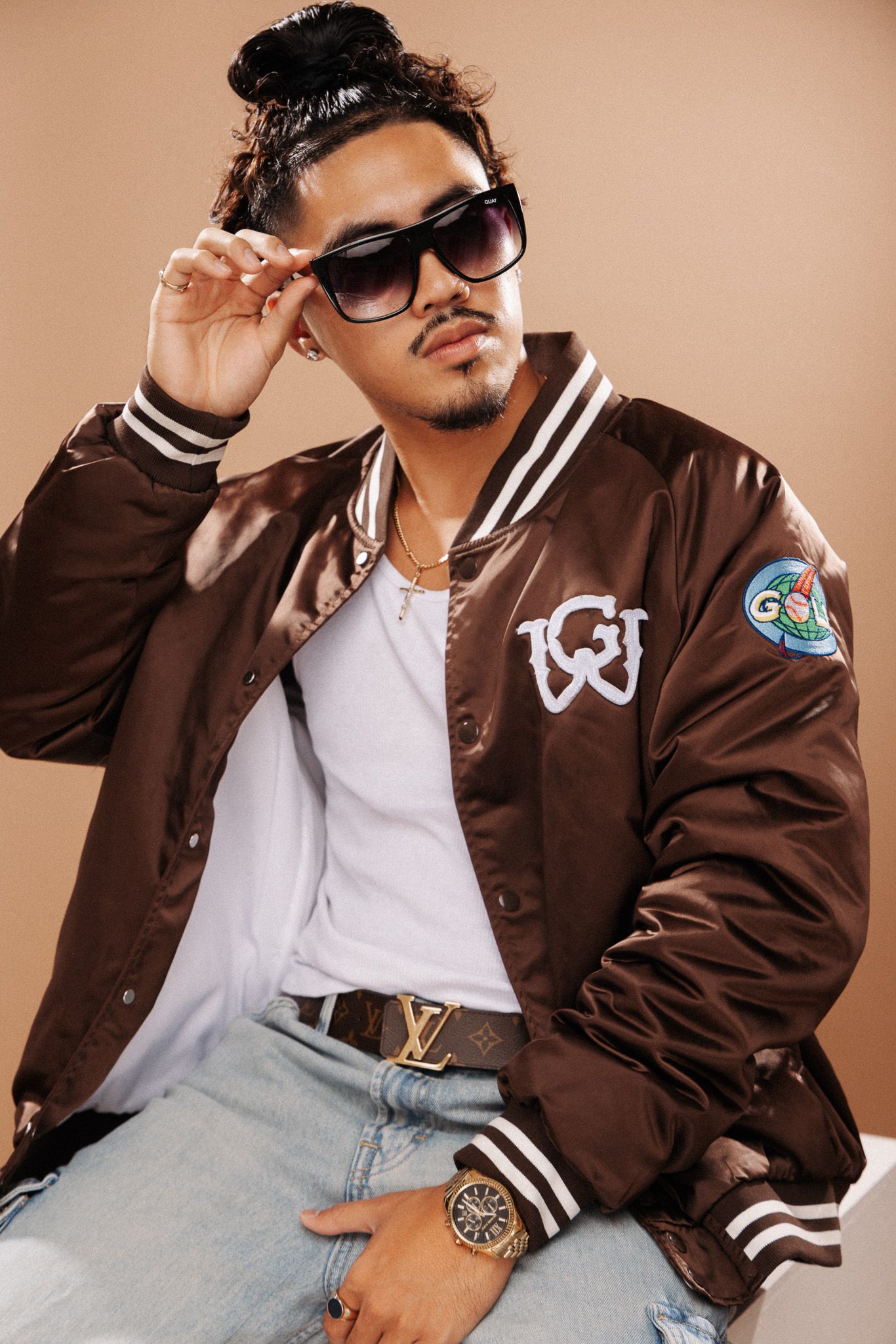 Model poses in brown varsity jacket with white stripes, Louis Vuitton belt, and sunglasses on beige background.