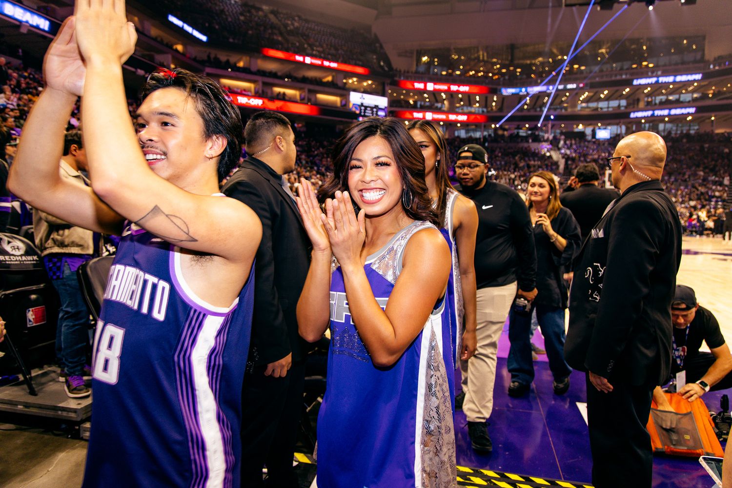 Excited fans celebrate courtside at Sacramento Kings NBA basketball game wearing purple team jerseys.
