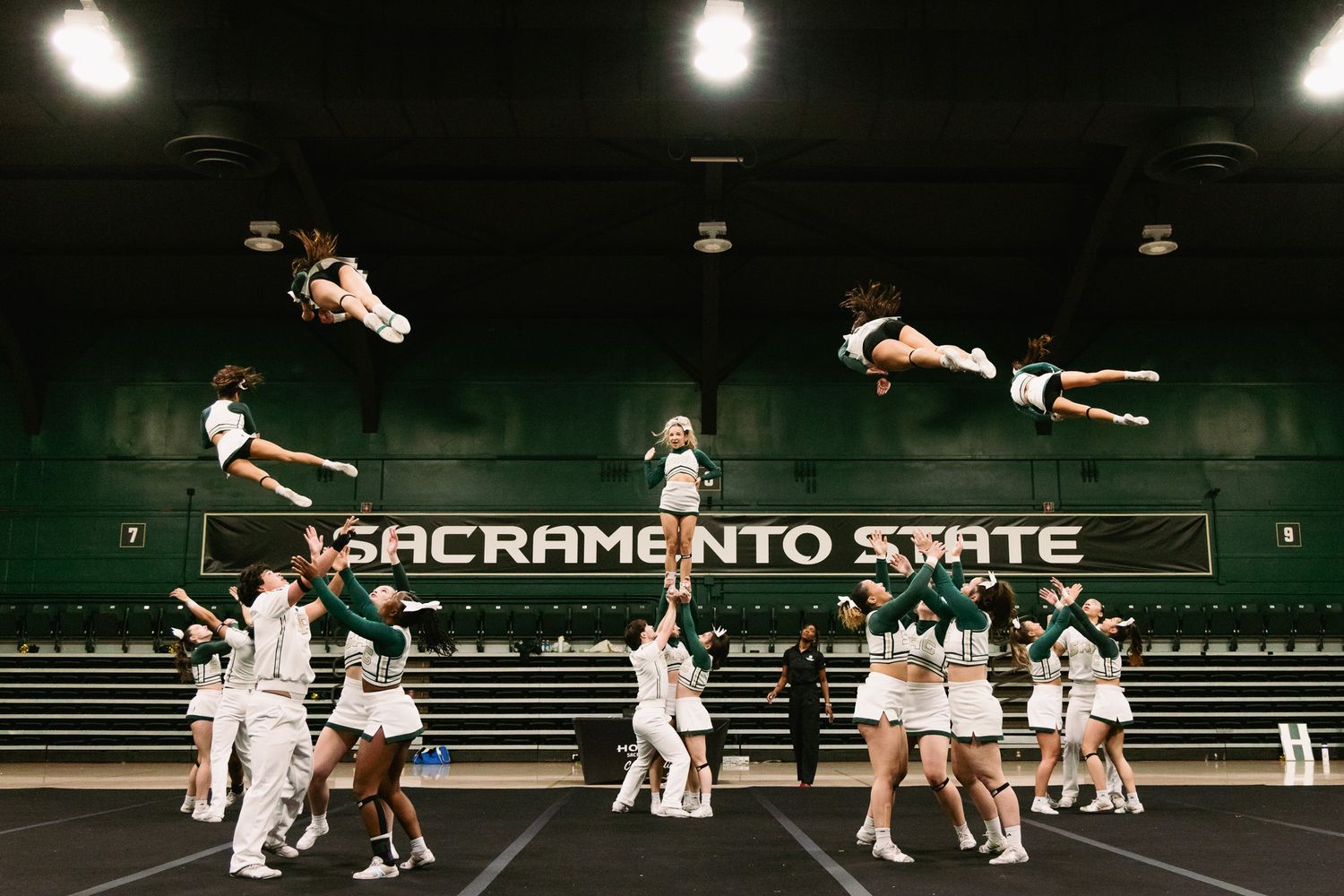 Sacramento State cheerleaders performing stunts with flyers soaring through the air during an indoor cheer routine.