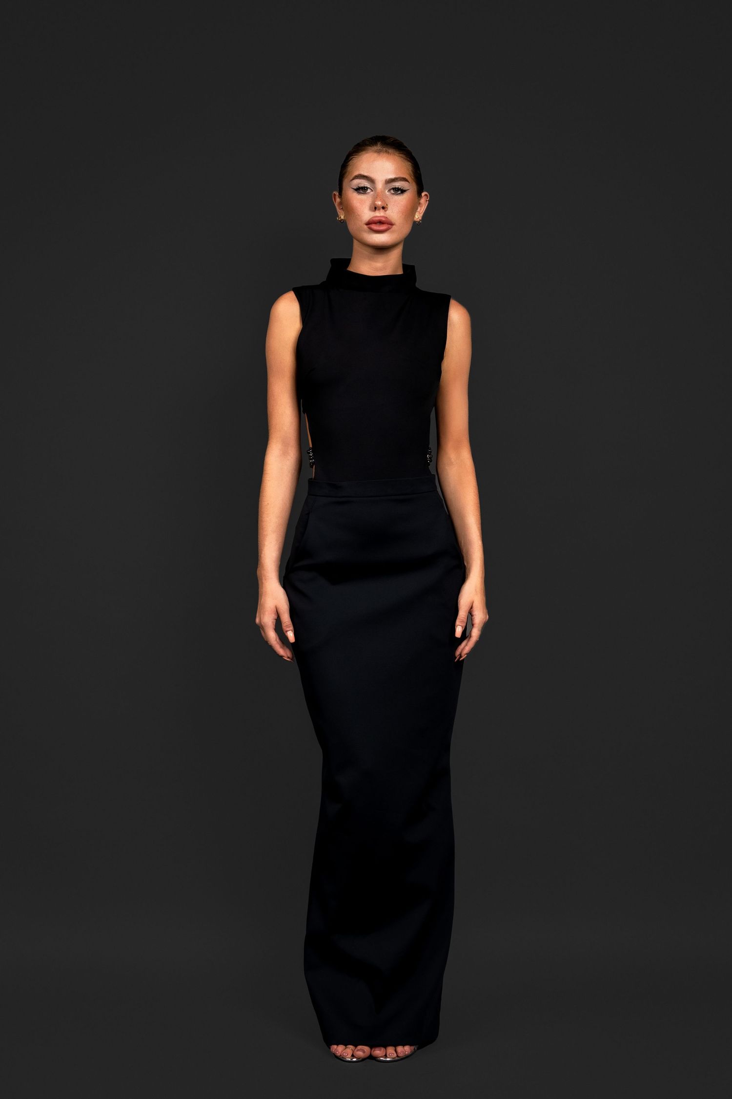 Model in a sleek all-black sleeveless turtleneck bodysuit and structured floor-length maxi skirt with gold earrings.