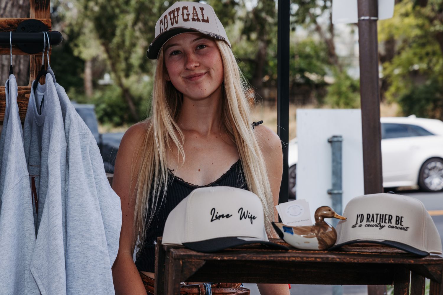 Smiling blonde woman wearing a cap displaying hats on a wooden rack outdoors at a casual retail stand.