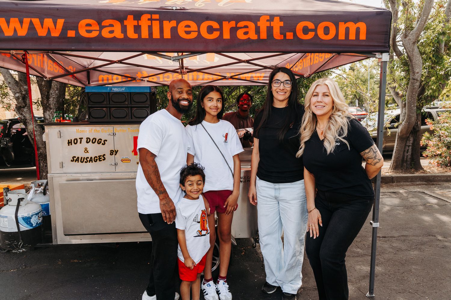Group of five people smiling in front of an Eat Fire Craft hot dogs and sausages food truck outdoors.