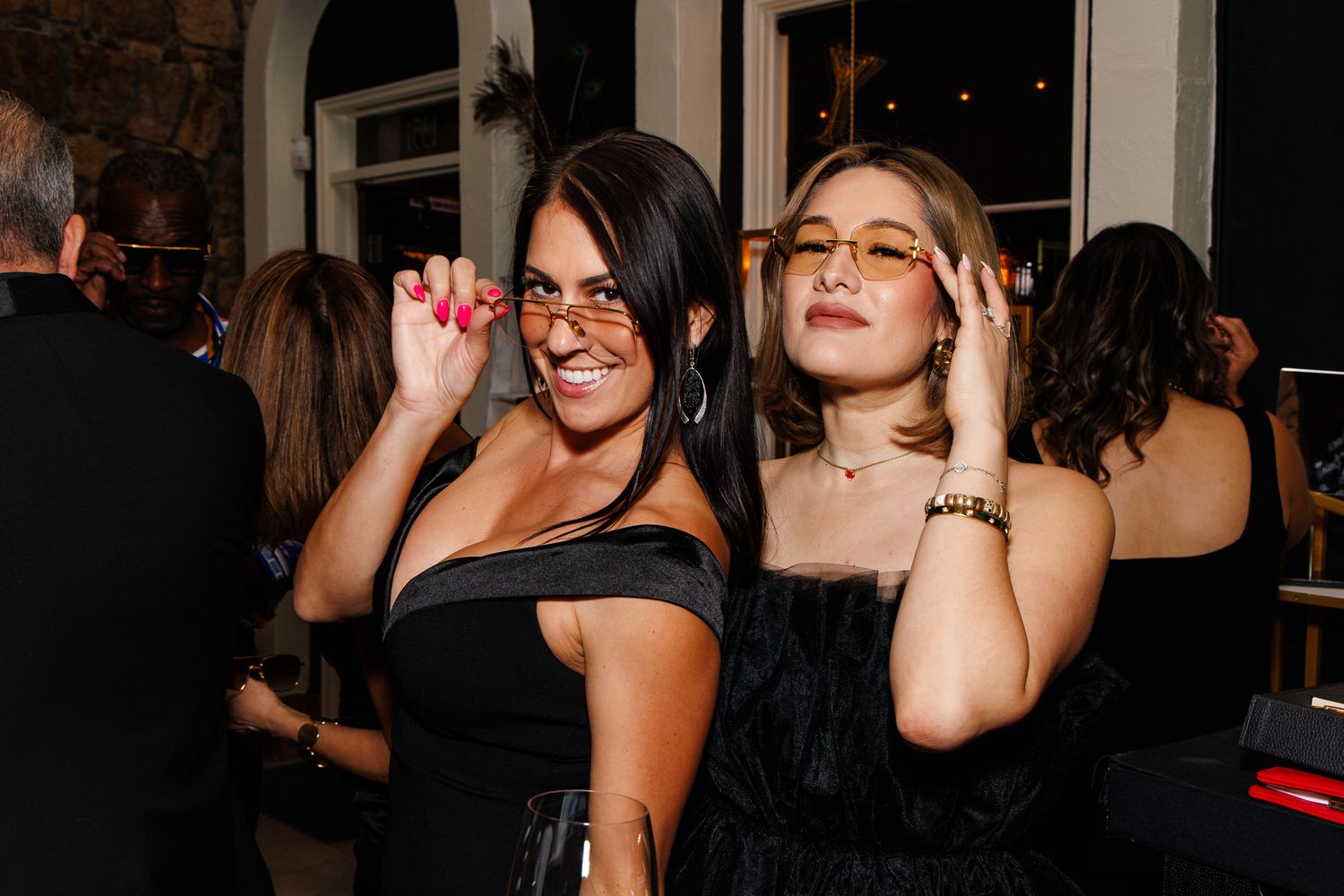 Two women in black outfits pose playfully wearing stylish tinted glasses at an evening event.