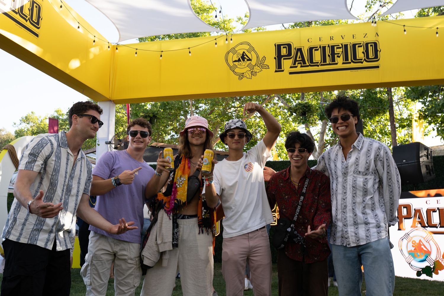 Group of people gathered outdoors under yellow tent canopies at what appears to be a Pacifico beer branded event.