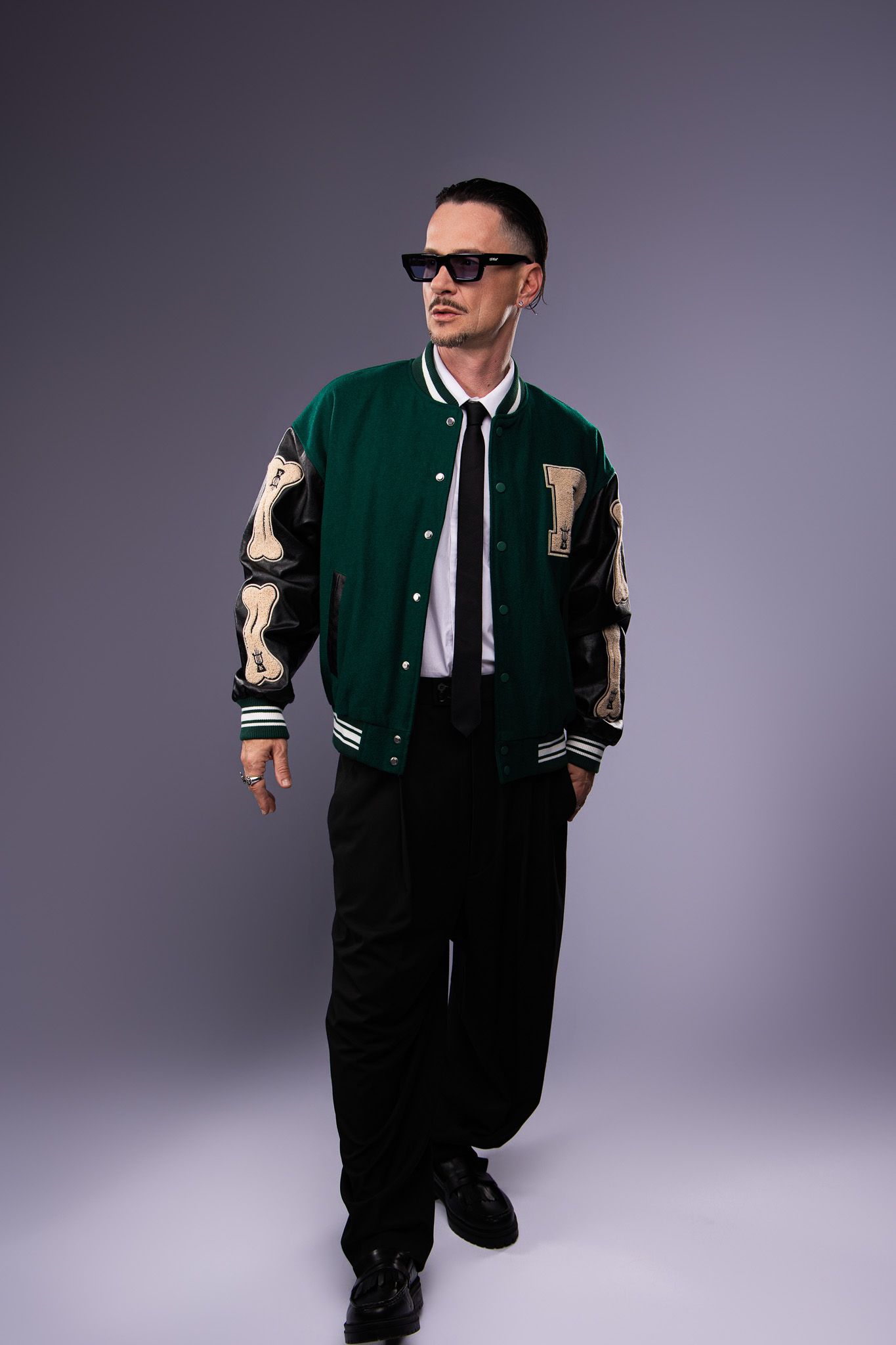 Varsity letterman jacket in green and cream worn with sunglasses, white shirt, tie and black pants against gray backdrop.