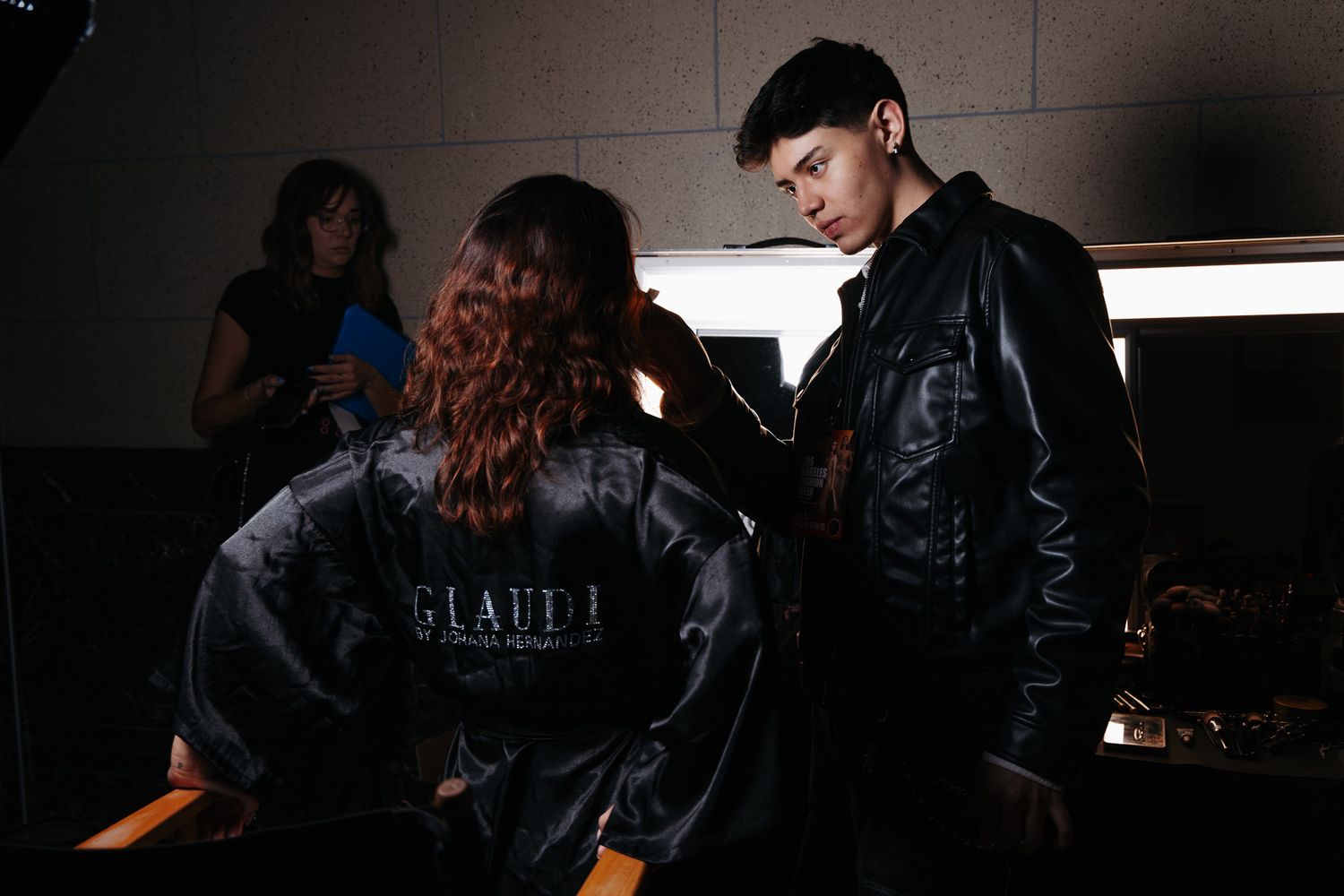 Dramatic black and white backstage scene with people in dark clothing and moody lighting.