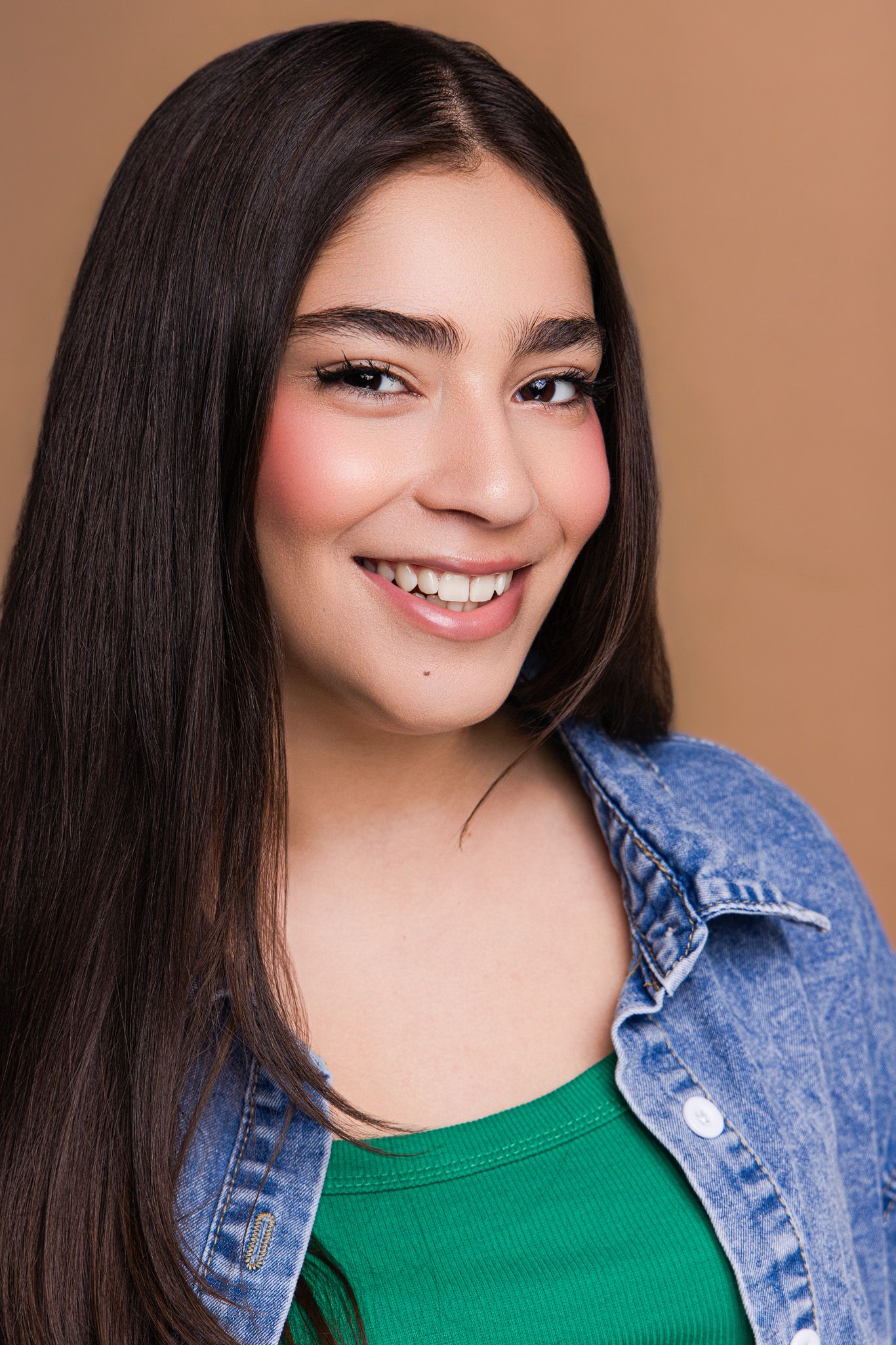 Smiling young woman with long dark hair wearing a green top and denim jacket against a beige background.