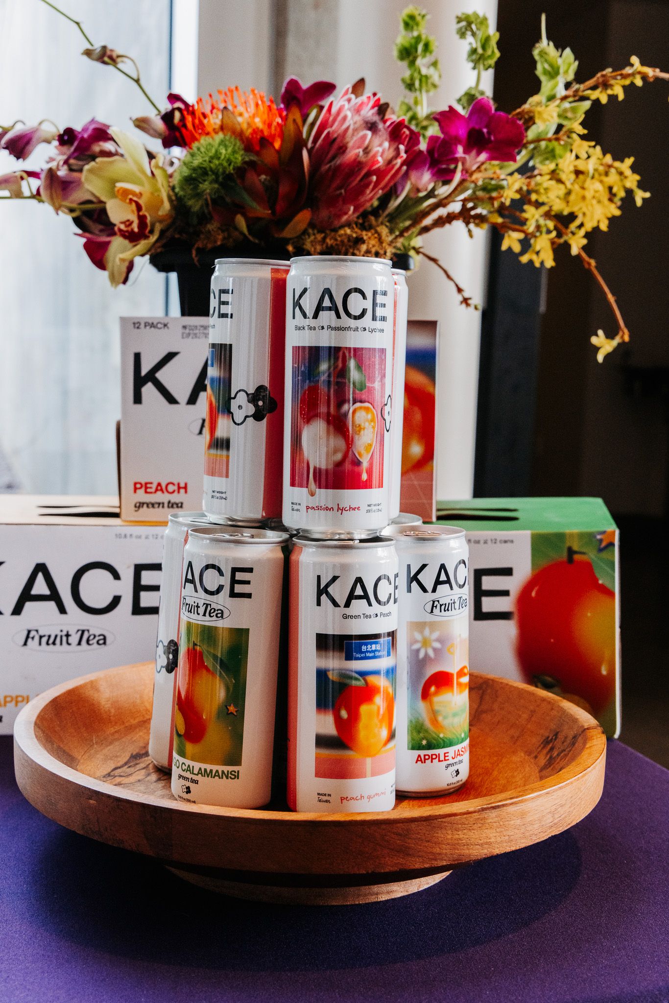 Colorful canned beverages and floral arrangements displayed on wooden circular tray with KACE brand products.
