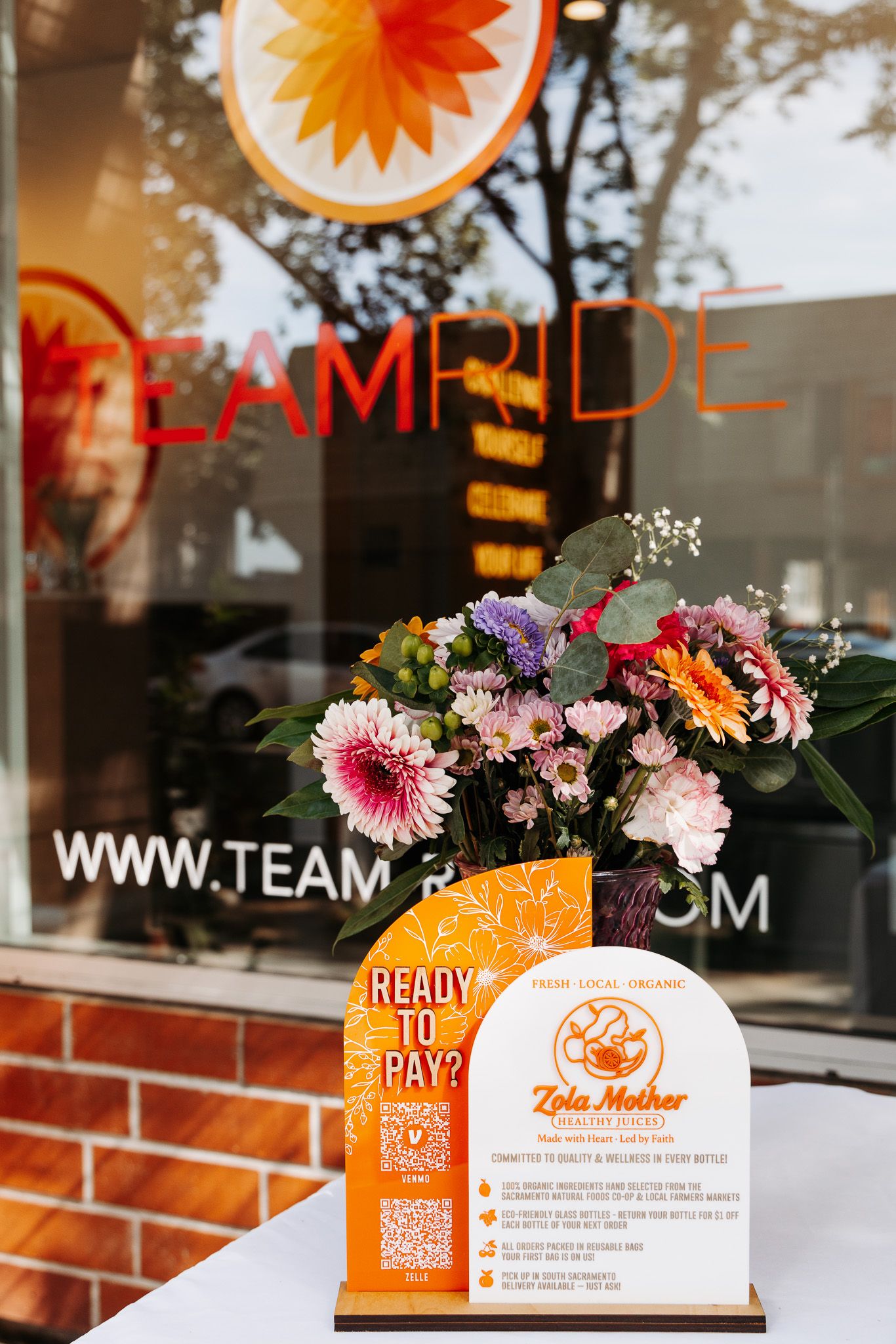 Business storefront window display with orange branding and pink floral arrangements on a brick exterior.