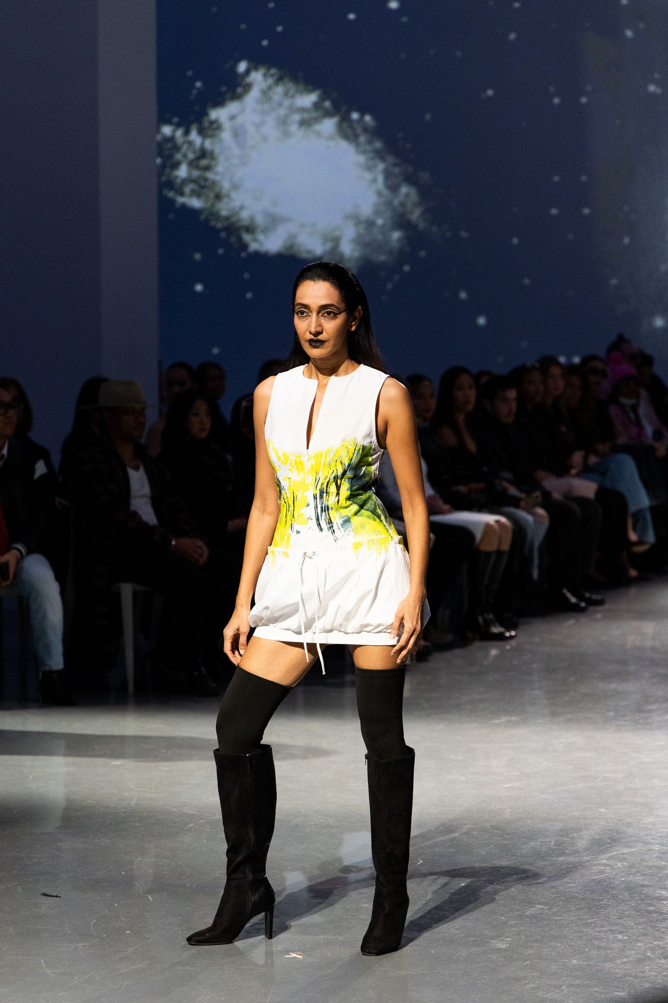 Model walks runway in white tank top and black pants against dark backdrop with audience.