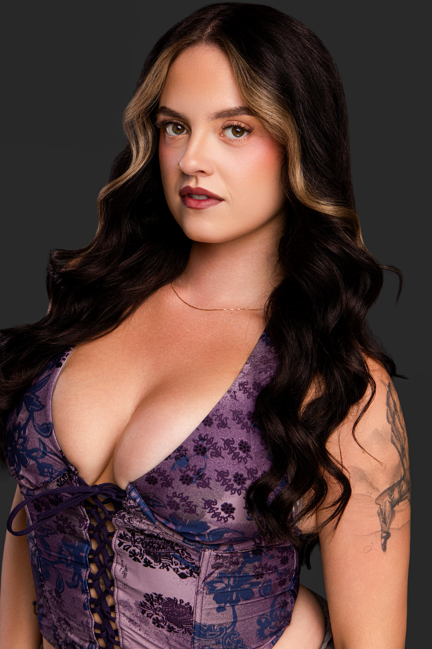 Woman with long dark wavy hair wearing a purple floral corset top, posing against a dark gray background.