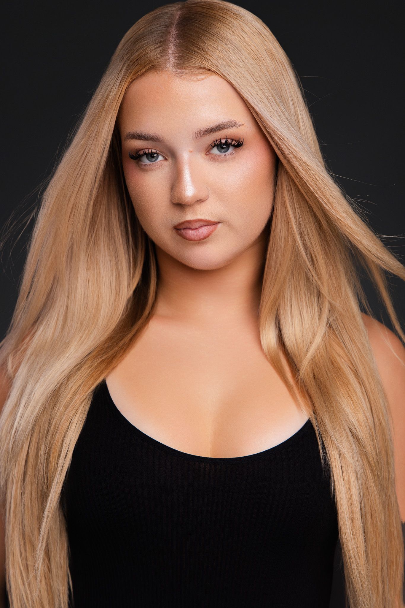 Young woman with long blonde hair wearing a black top, posing against a dark background in a professional portrait.