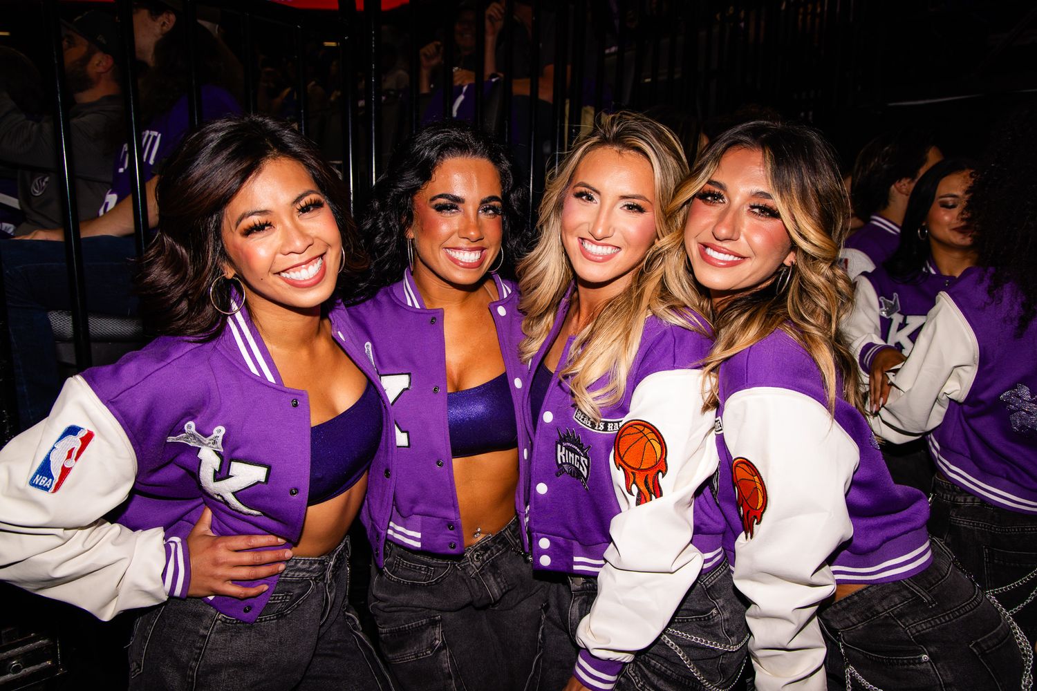 Four women in purple NBA varsity jackets pose together smiling at a nightclub event.
