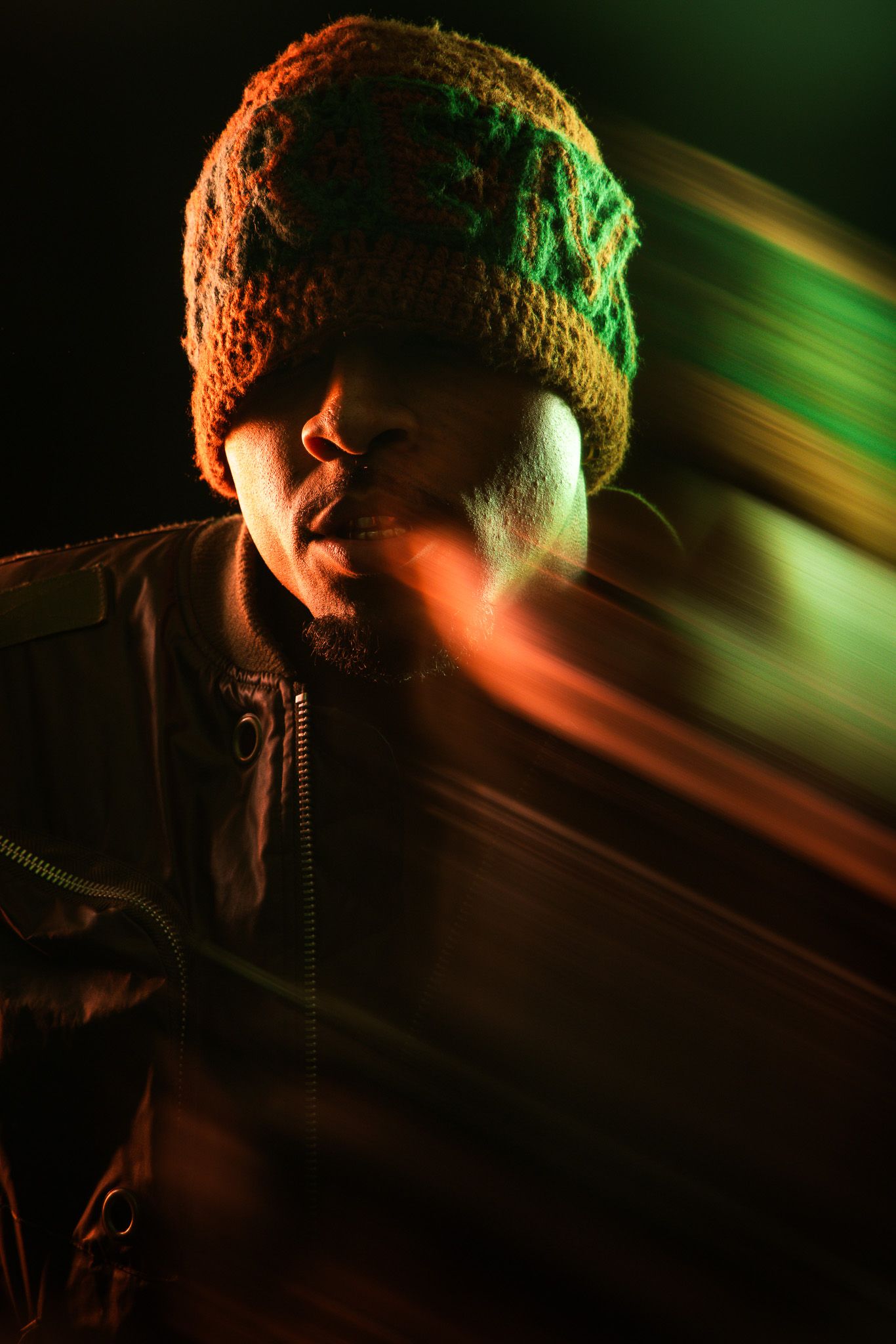 Man in colorful knit beanie and brown jacket posed with dynamic light streaks in dark studio.