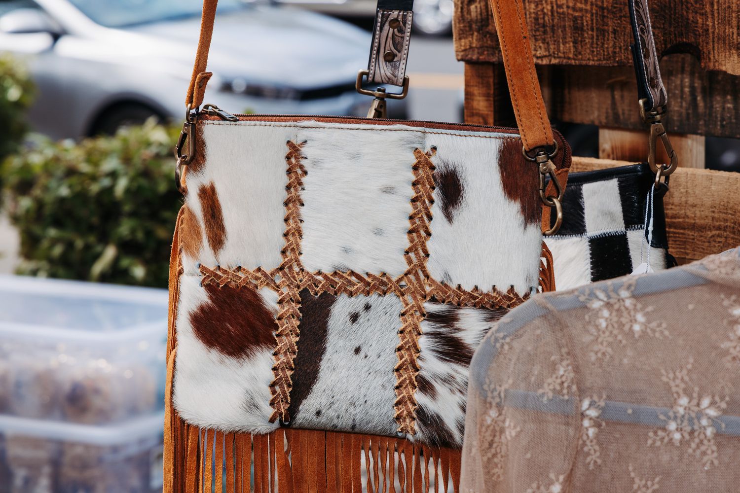 Cowhide leather crossbody bag with fringe and whipstitch detailing displayed at an outdoor market.