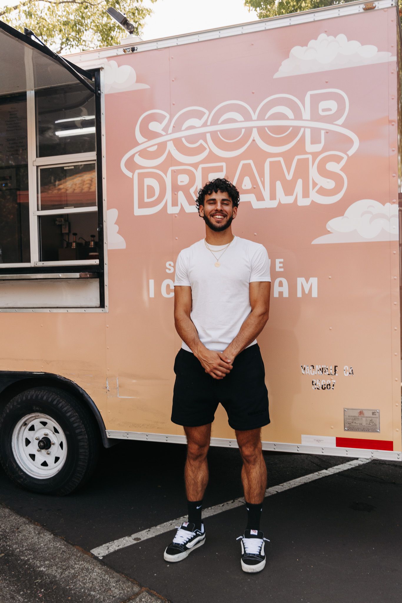 A young man in a white t-shirt and black shorts stands smiling in front of a Scoop Dreams ice cream food truck.