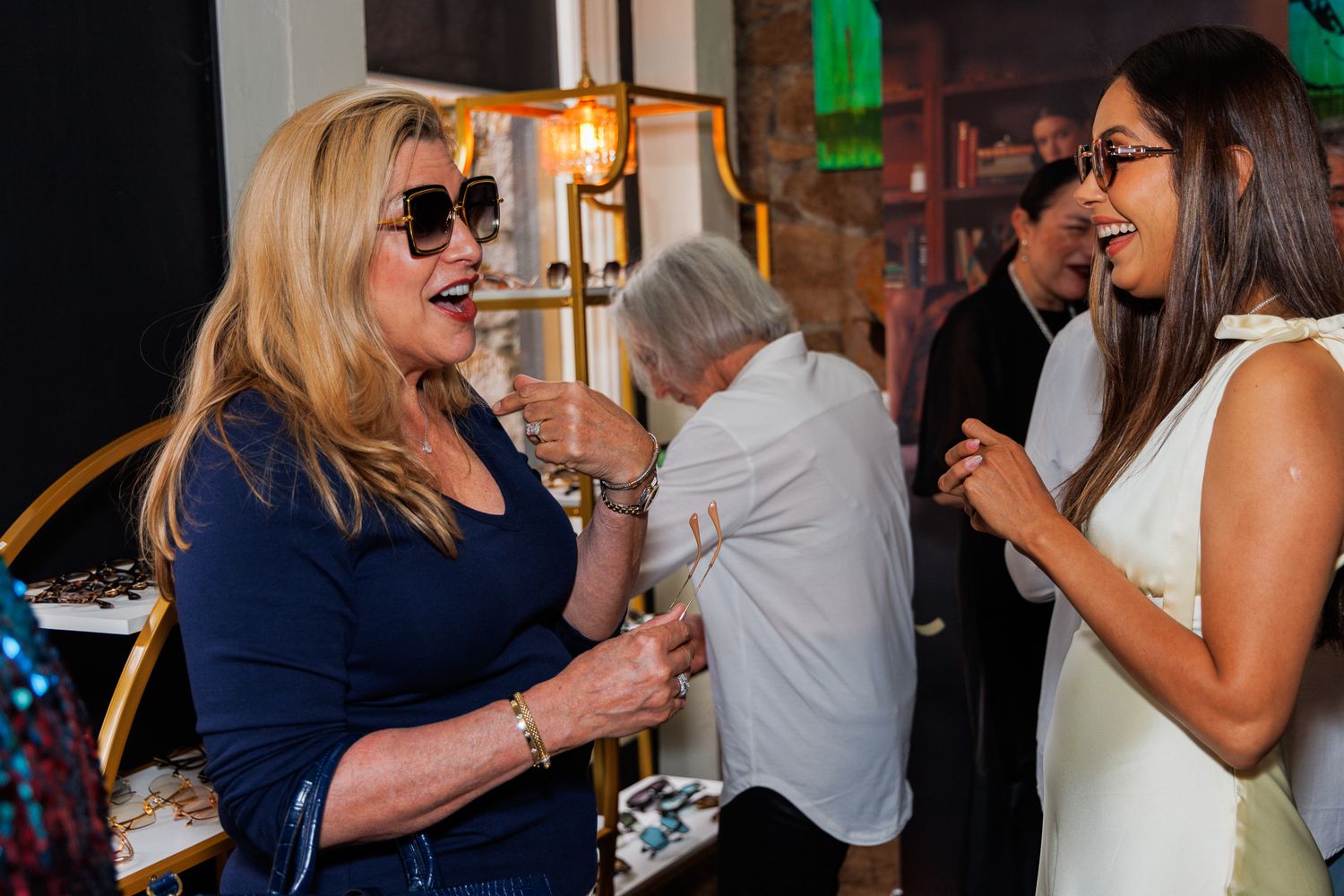 Two women laughing and socializing at an indoor event, one wearing sunglasses and blue top, other in white dress.