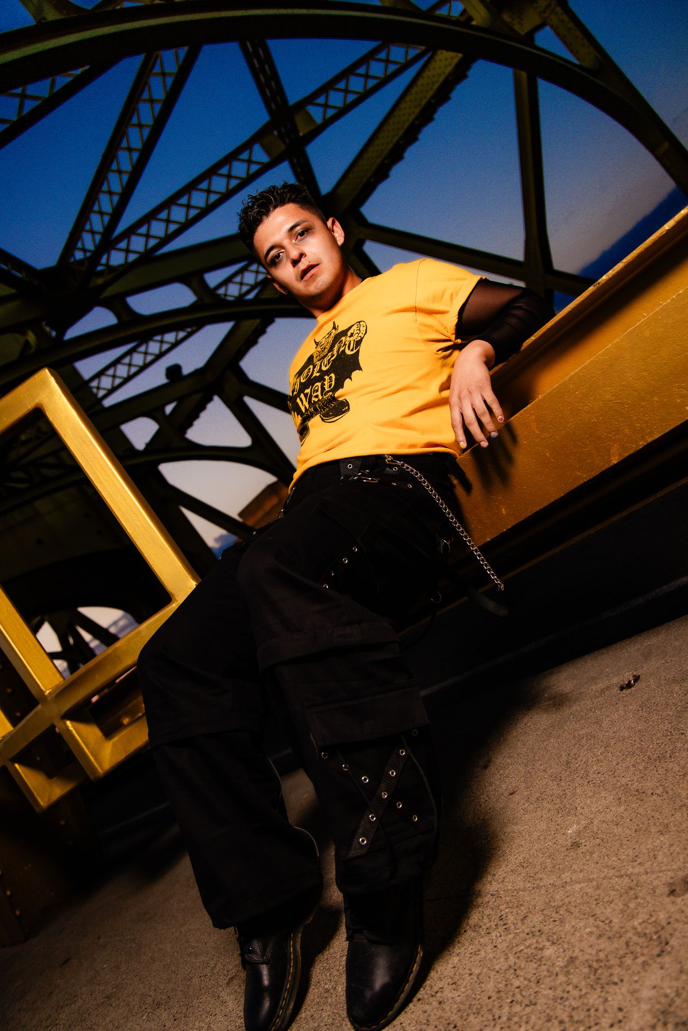 Person in yellow shirt sitting against industrial bridge structure under dramatic lighting with geometric shadows.