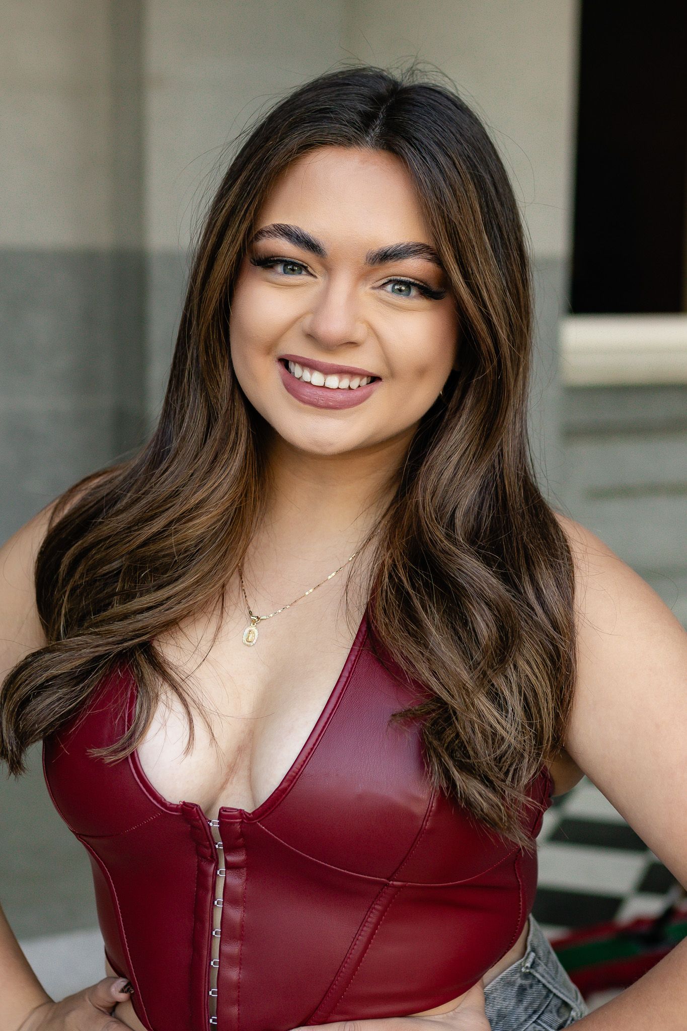 A person poses in a burgundy leather top against a light-colored architectural background.