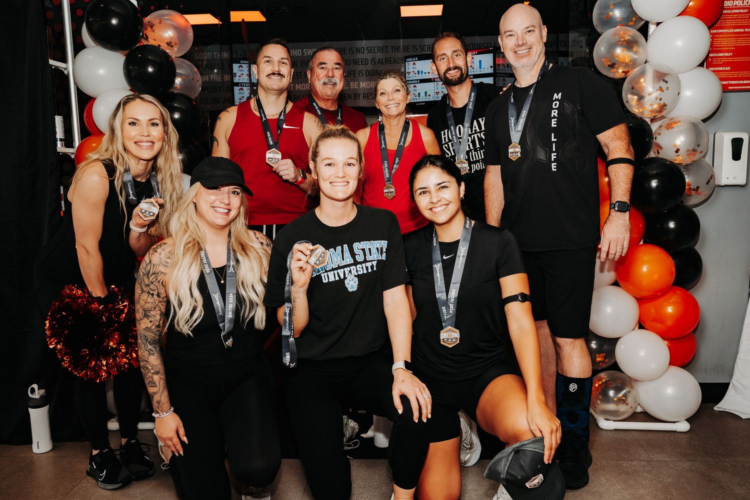 Group of people posing together at a party decorated with orange and white balloons in a gym setting.