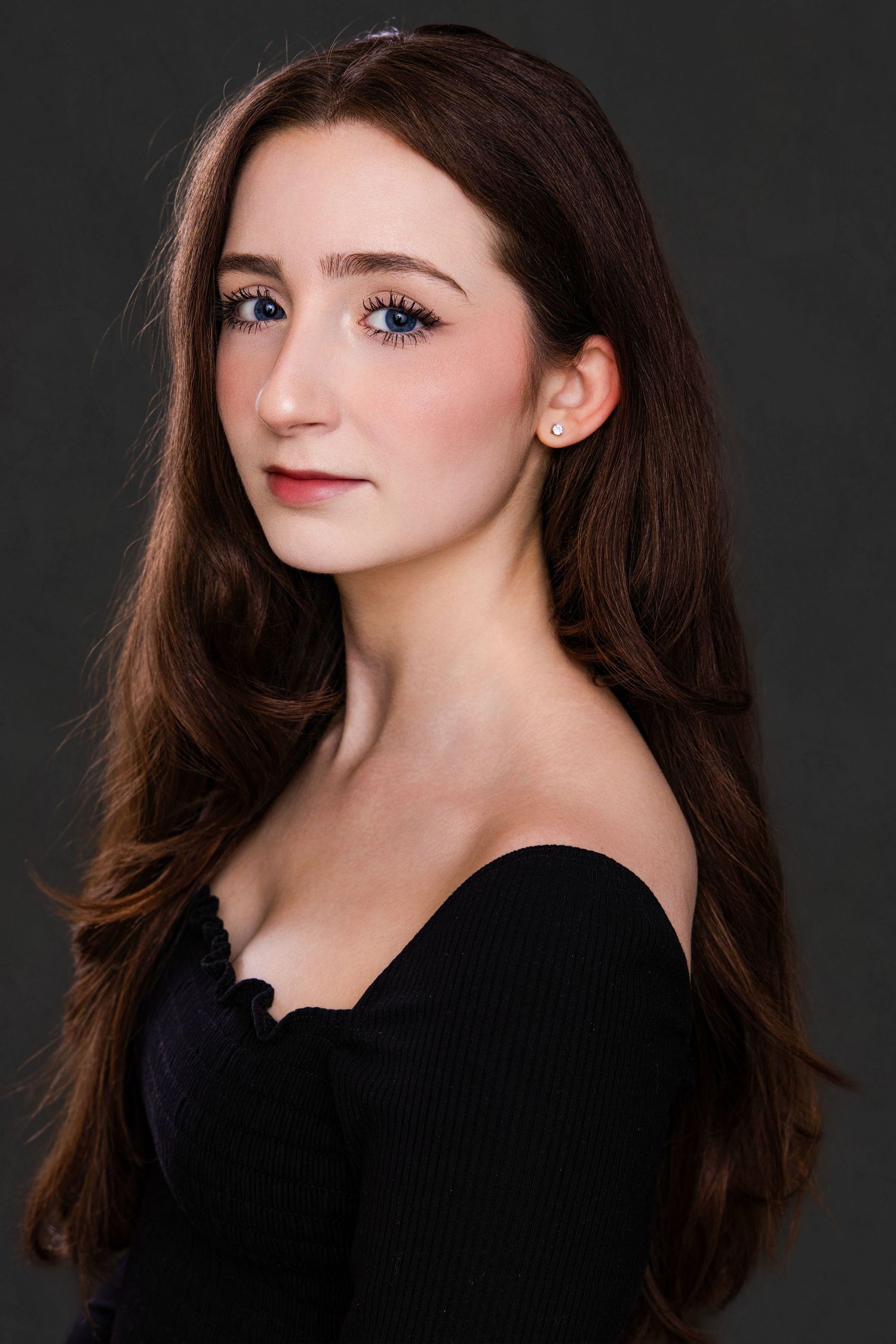 Young woman with long brown hair and blue eyes wearing a black off-shoulder top, posing for a professional portrait.