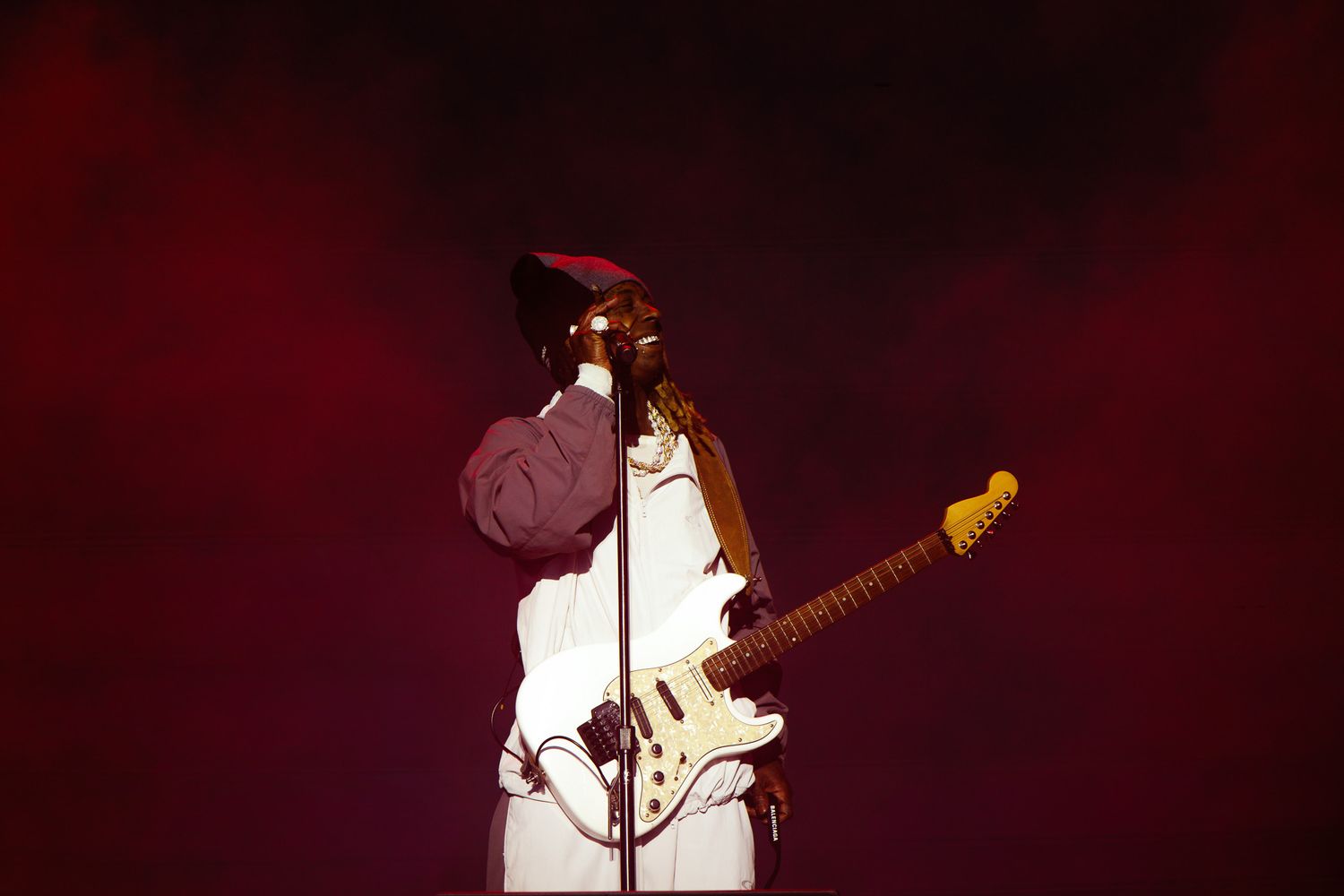 Musician playing white Fender guitar on stage under dramatic red and pink lighting during live concert performance.