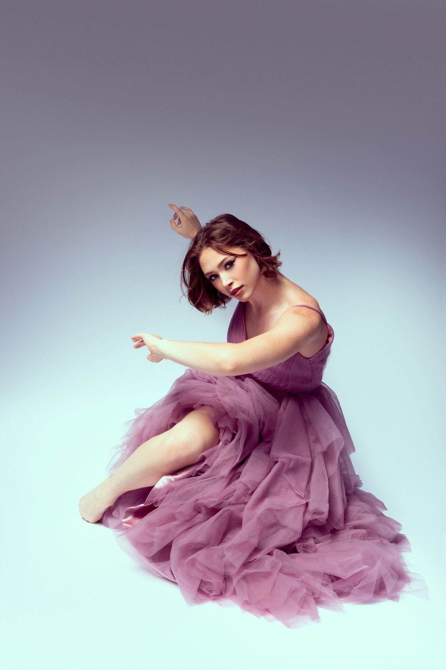 A dancer in a flowing mauve tulle dress performs graceful movements against a light studio backdrop.