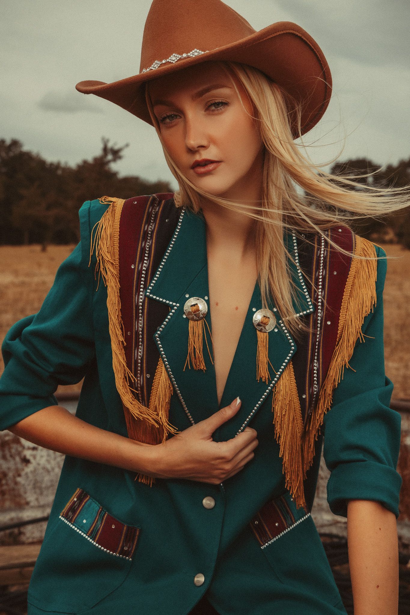 Western-style fashion portrait featuring teal fringe jacket with gold tassels and brown leather cowboy hat in rustic setting.