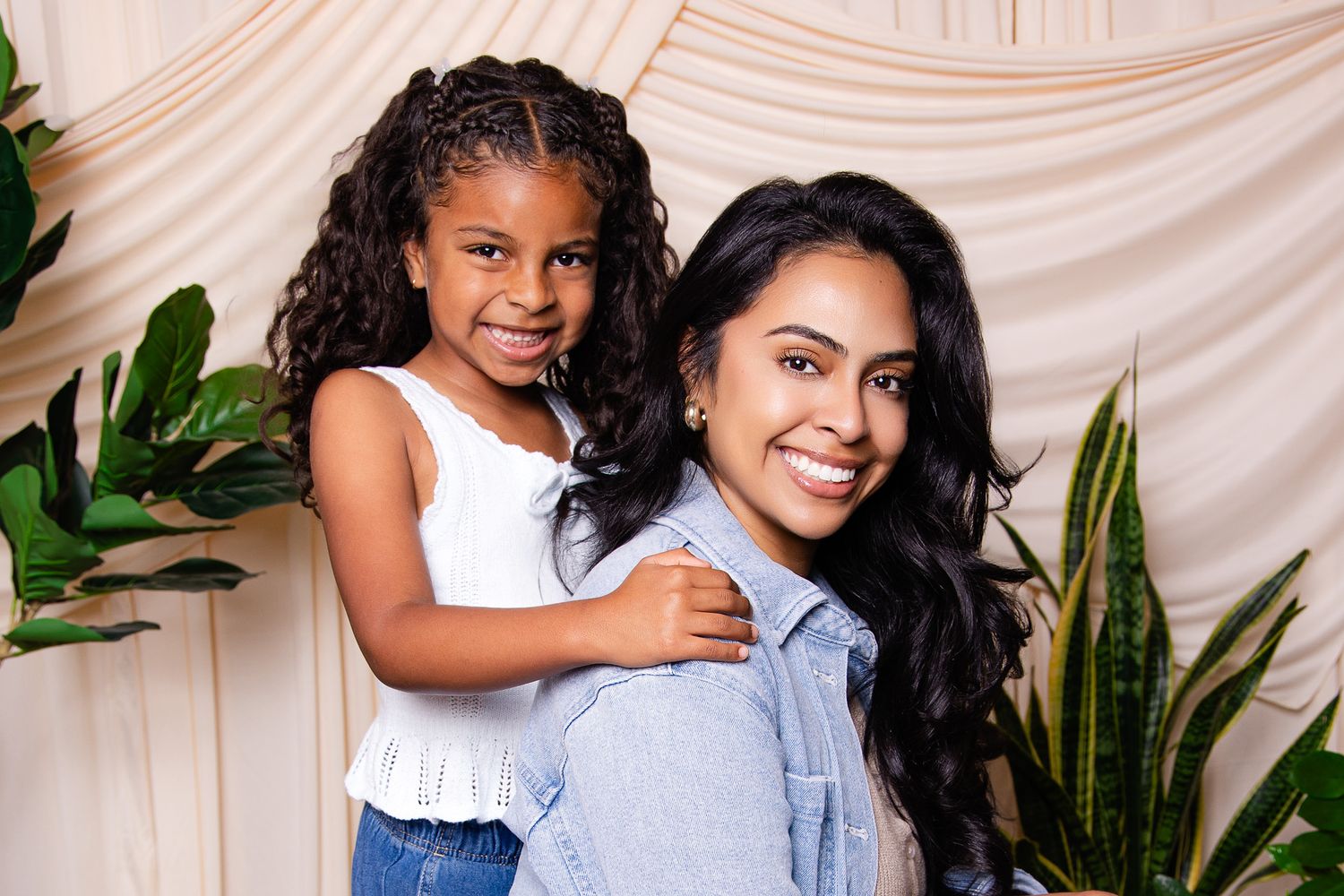 A woman and young girl smile together in front of a neutral backdrop with green plants.