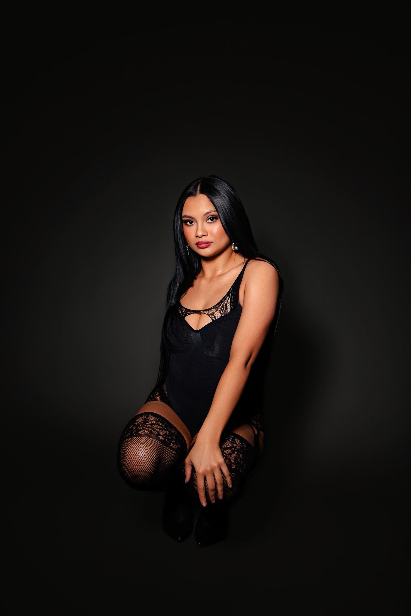 Woman in black lingerie and lace stockings posing on dark background in a professional boudoir photo.