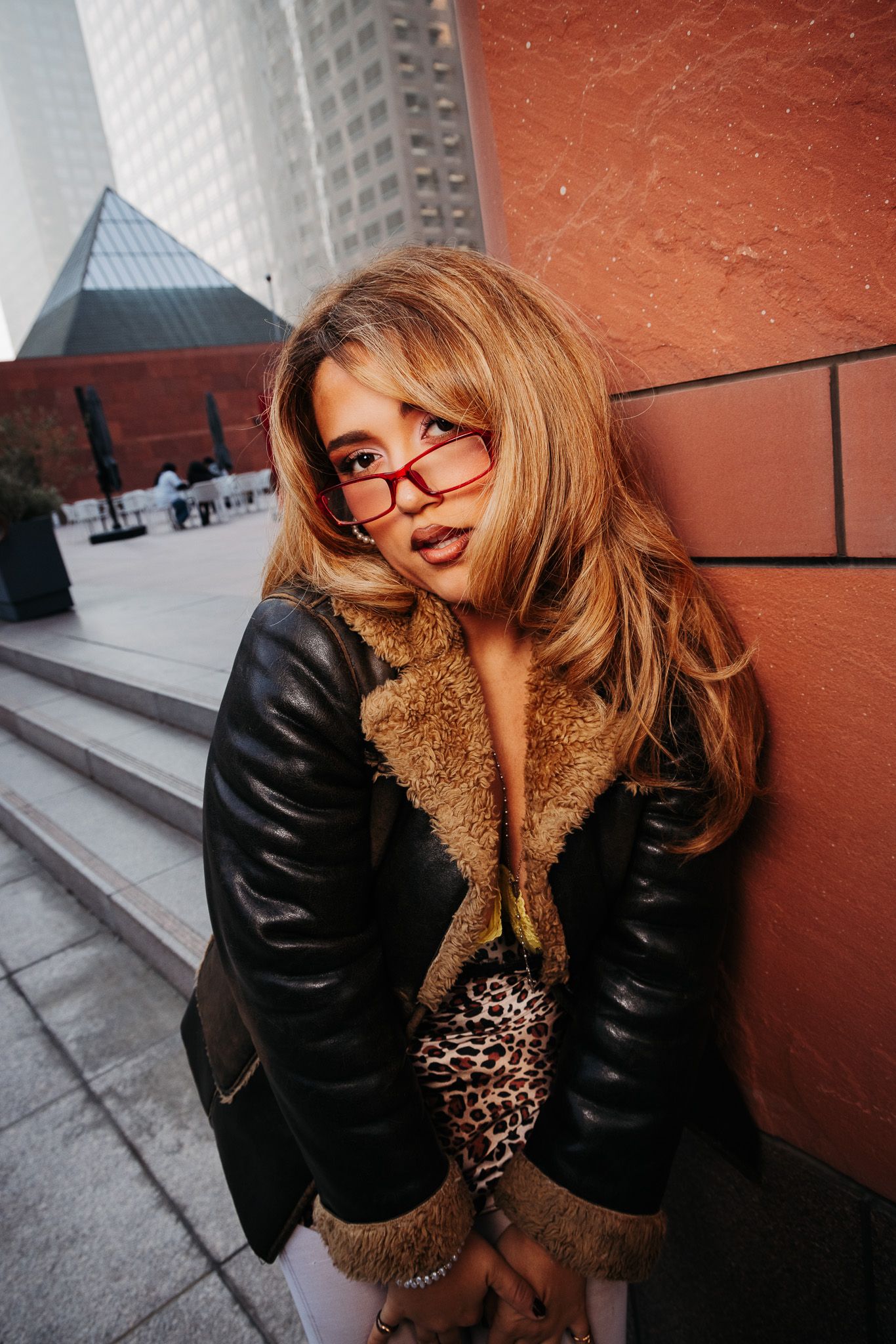 Person in leather jacket with fur collar and glasses poses against red wall in urban setting.