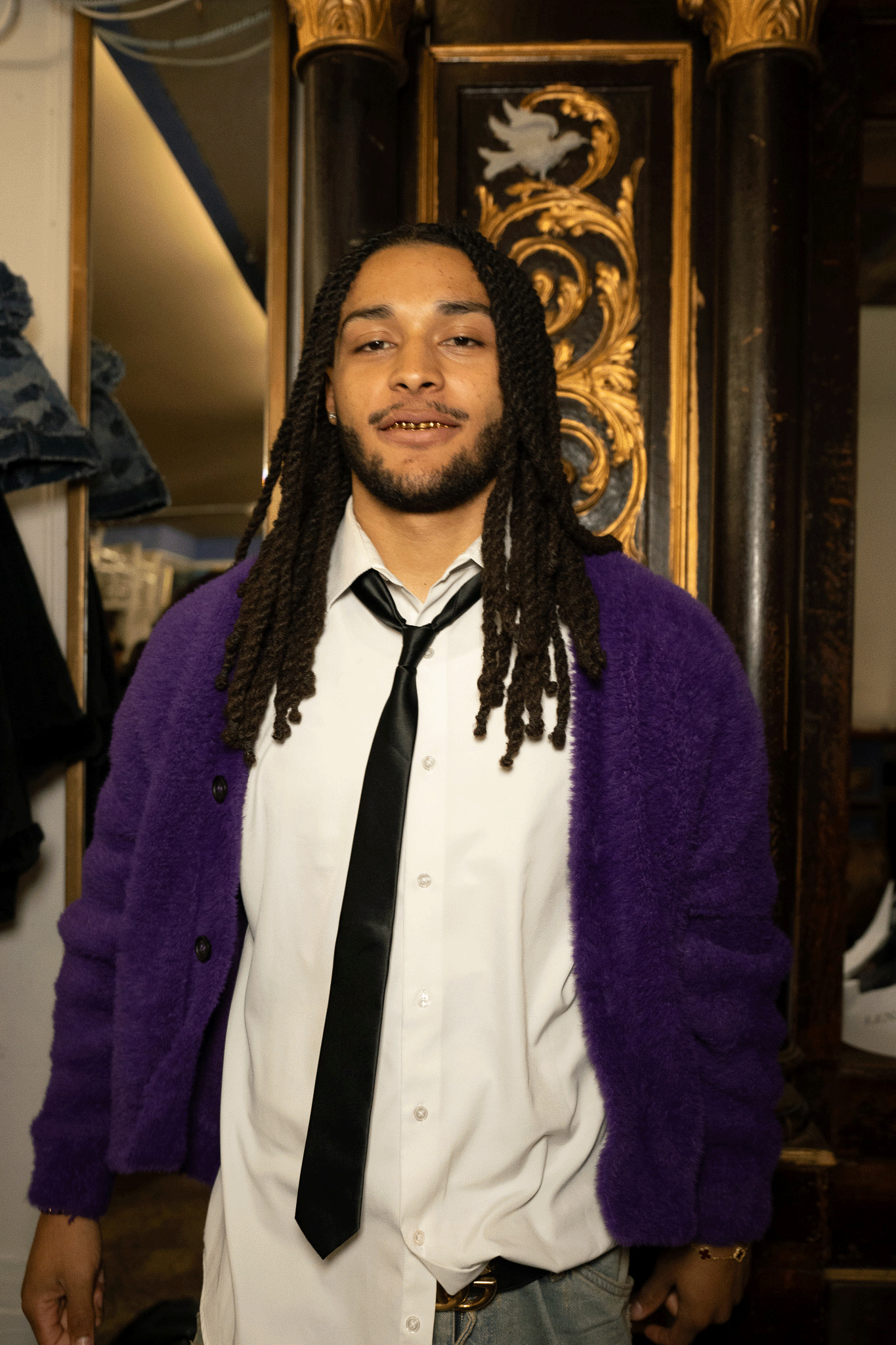 Person wearing a purple cardigan, white dress shirt and black tie smiling at the camera in an indoor setting.