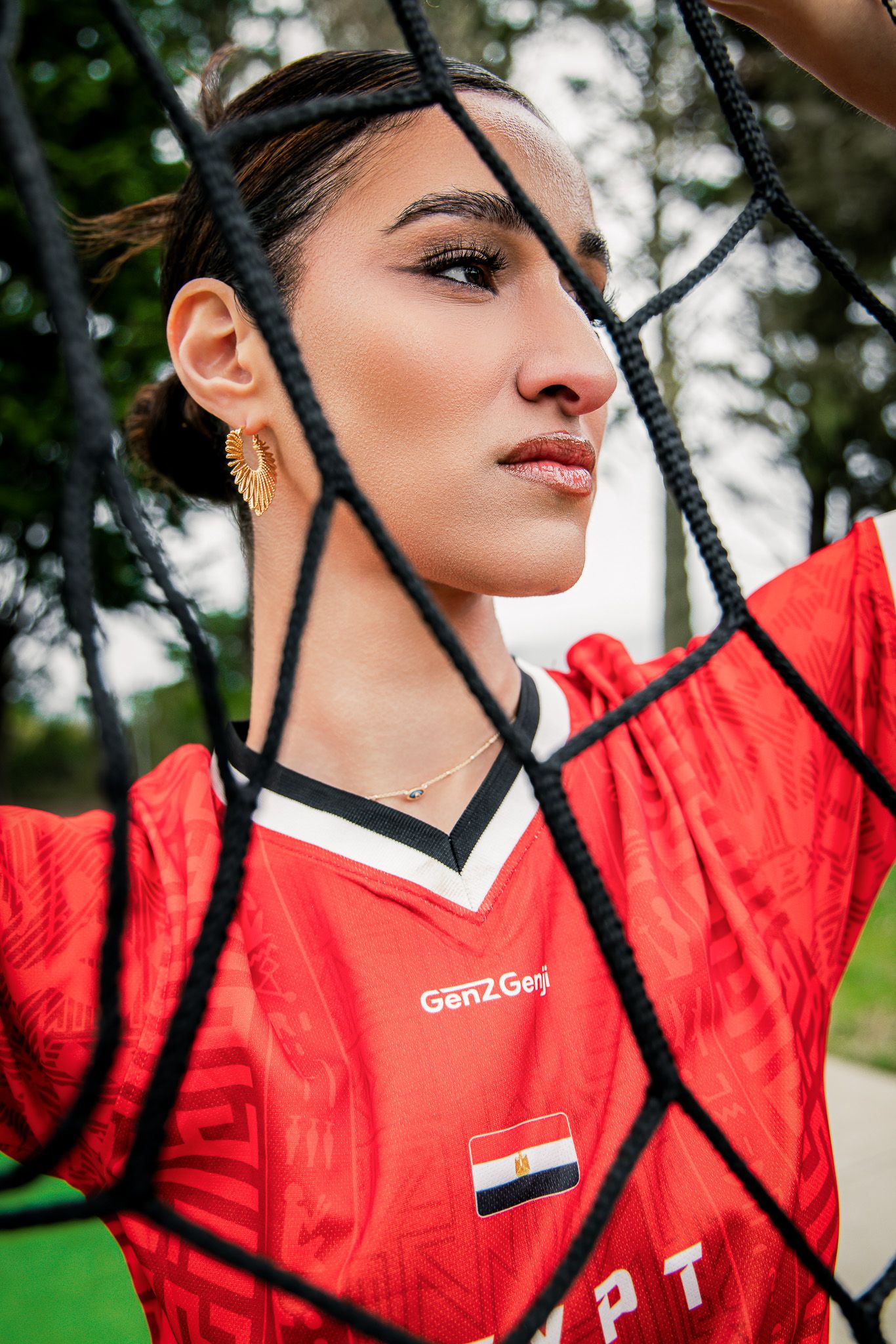 Young woman in red Egypt soccer jersey poses behind a black net outdoors, wearing gold earrings and necklace.