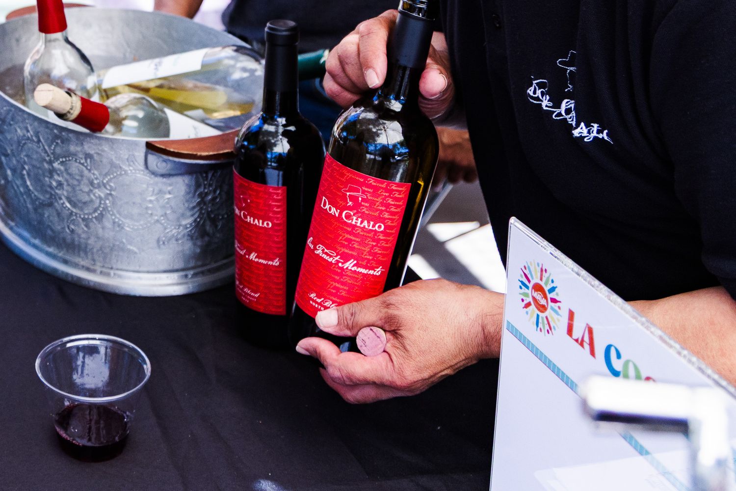 Person holding two red-labeled Don Camilo wine bottles at an outdoor tasting event with ice bucket.