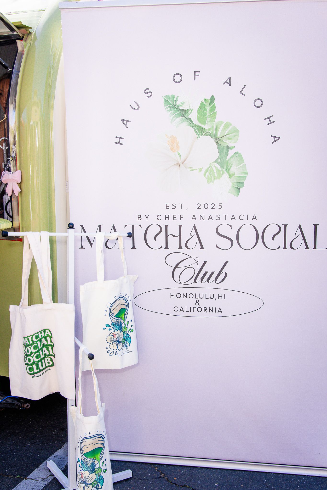 A white backdrop display for Matcha Social Club with green leaf logo and branded merchandise bags on a stand.