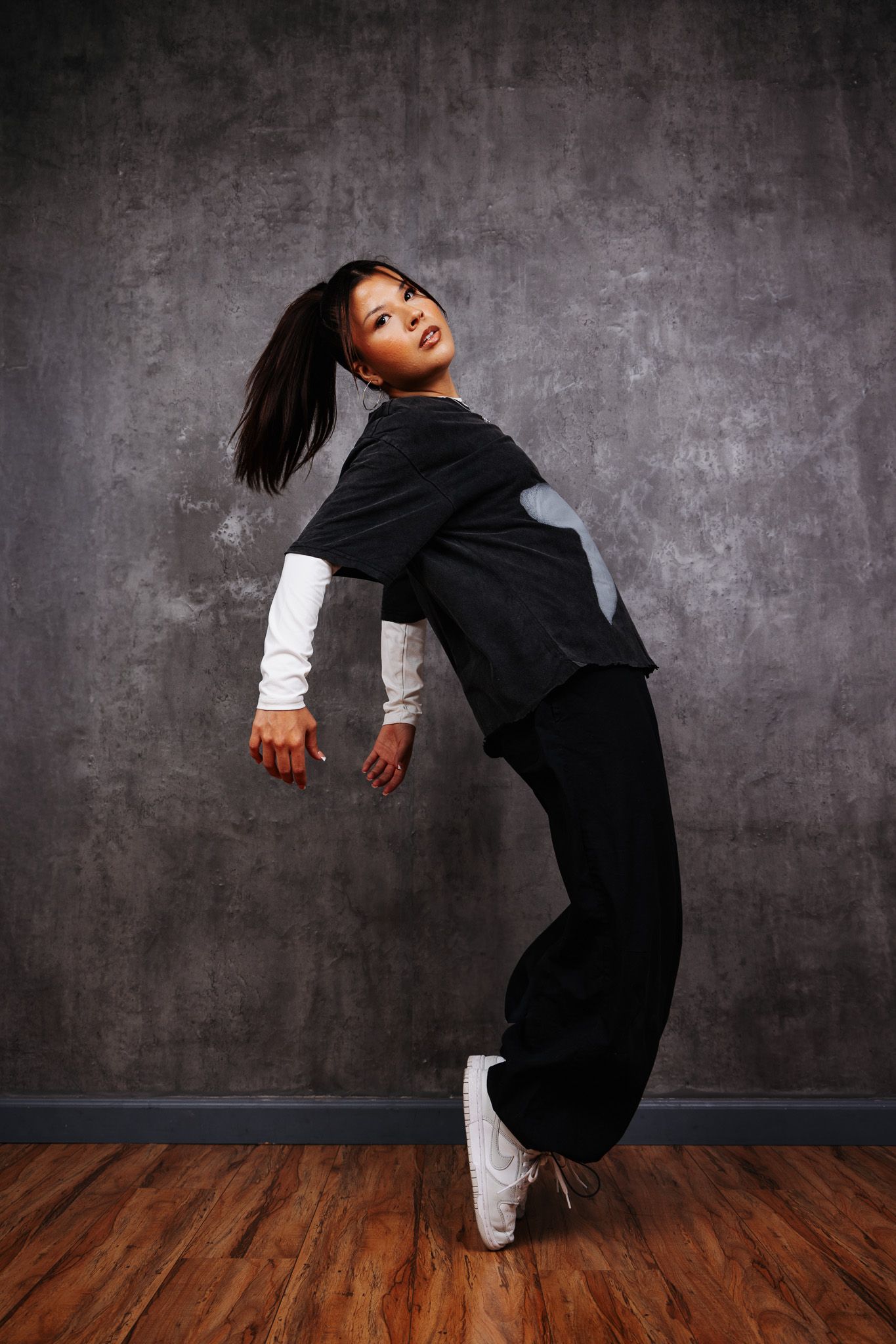 Dynamic dancer in black outfit with white sleeves performs a dramatic backbend pose against a textured gray wall.