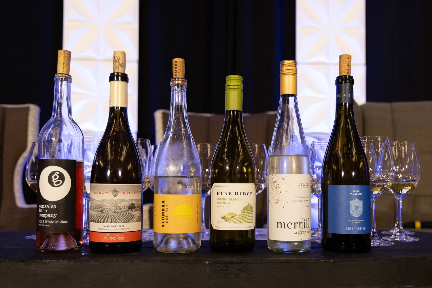 Six wine bottles of different varieties displayed on a dark surface with empty wine glasses.