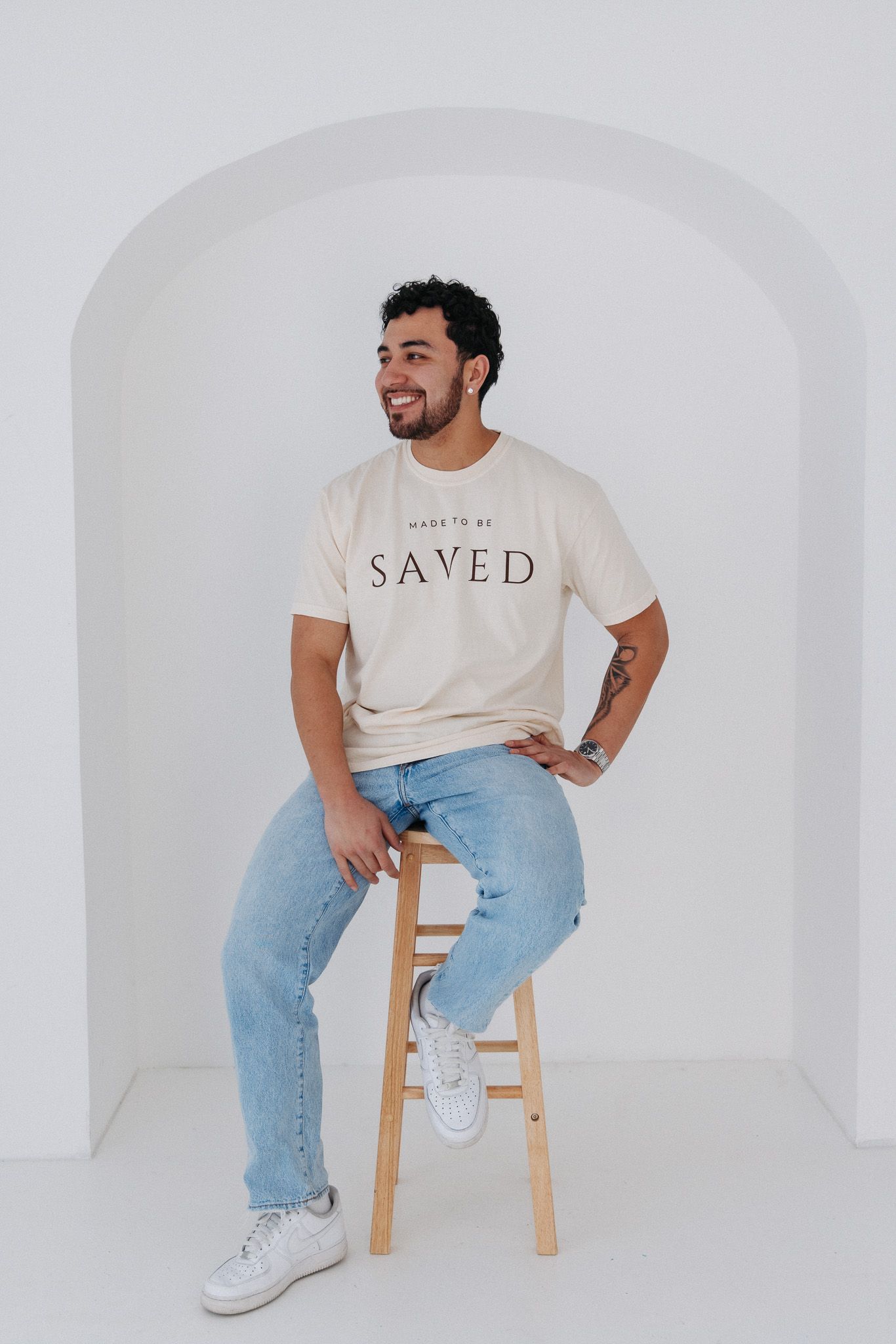 Casual portrait of person in white 'SAVED' t-shirt and jeans sitting on wooden stool against white wall.